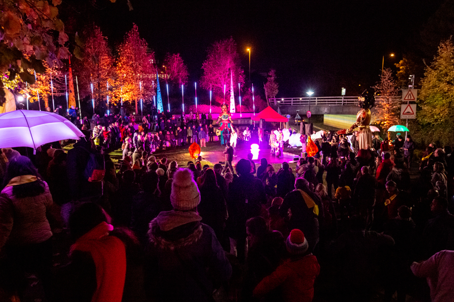 An outdoor event, crowds of people surround a performance. It is night time but strips of lights light up the performance space.