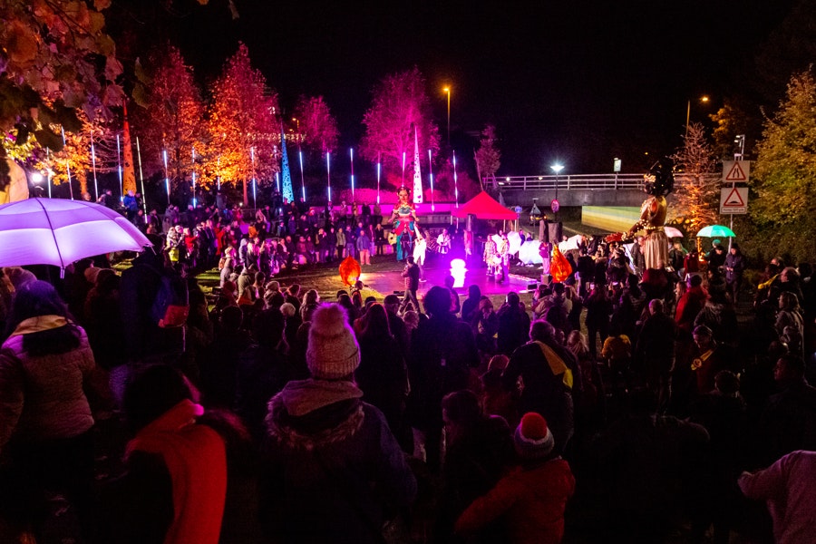 An outdoor event, crowds of people surround a performance. It is night time but strips of lights light up the performance space.