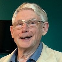A close up 'head and shoulders' photograph shows a man smiling at the camera. He wears a beige coat and blue shirt. He also wears light framed glasses.