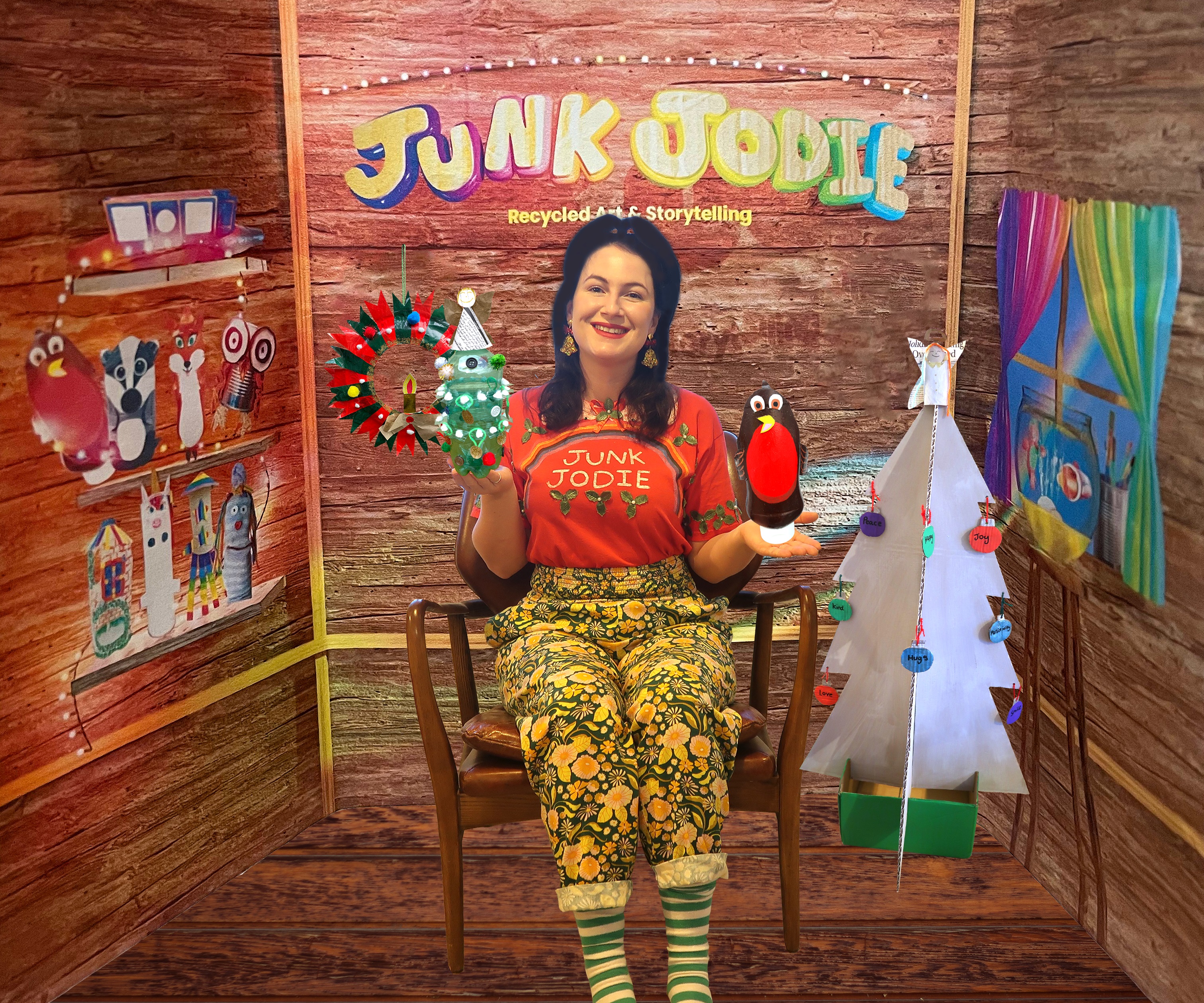 Junk Jodie sits on a wooden chair, holding a puppet robin and Christmas tree made from cardboard. Jodie wears a red tshirt and trousers covered in a gingerbread man design.