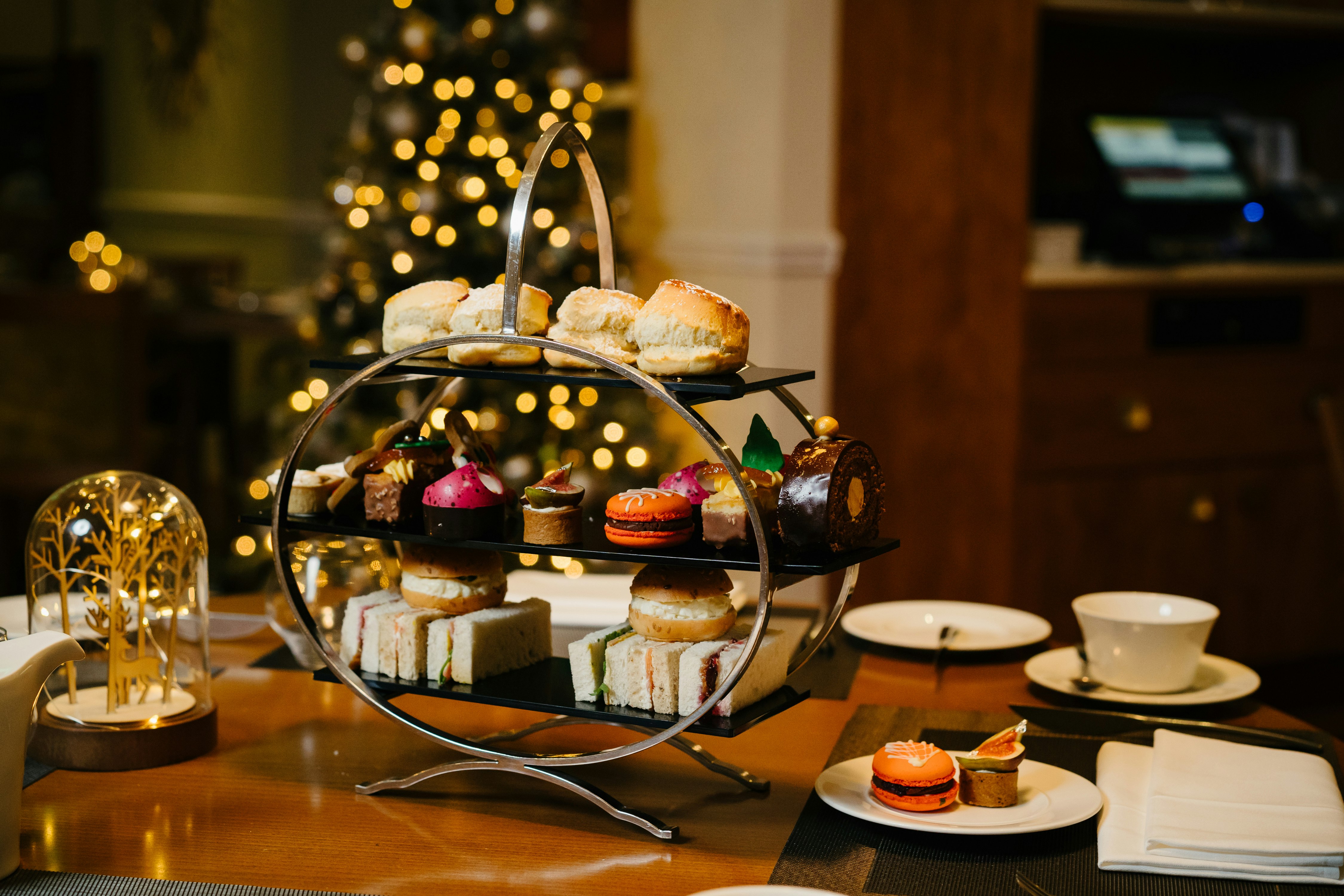 A silver cake stand sits on top of a wooden table, a Christmas tree with fairy lights twinkles in the background. There are cakes, scones and sandwiches stacked on the cake stand.