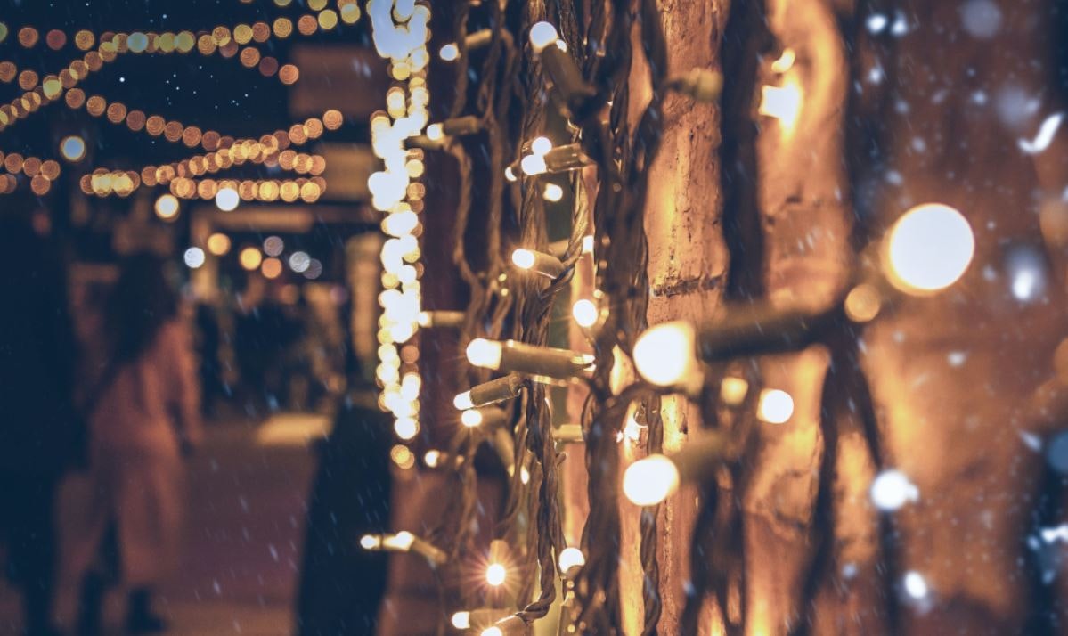 Twinkly fairy lights are pictured against a brick wall. Blurred in the background of the image people walk around in a Christmas fayre.