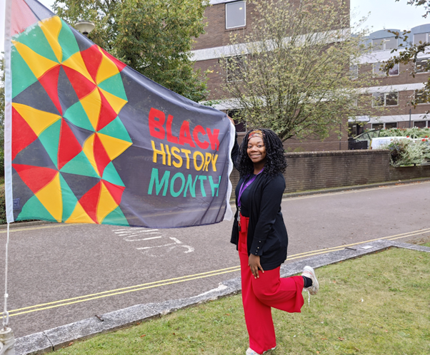 A person smiles at the camera holding a Black History Month flag. They wear red trousers and a black top and cardigan.