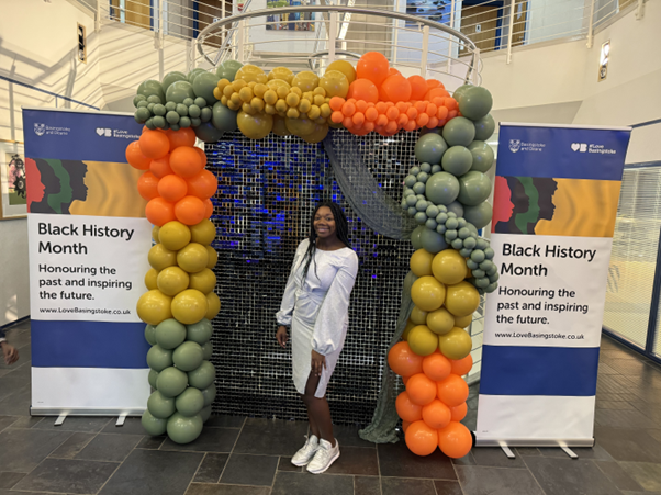 Jade smiles at the camera, she wears a white fitted knee length dress. Jade stands infront of a balloon display, either side of the display are two banners highlighting black history month celebrations.