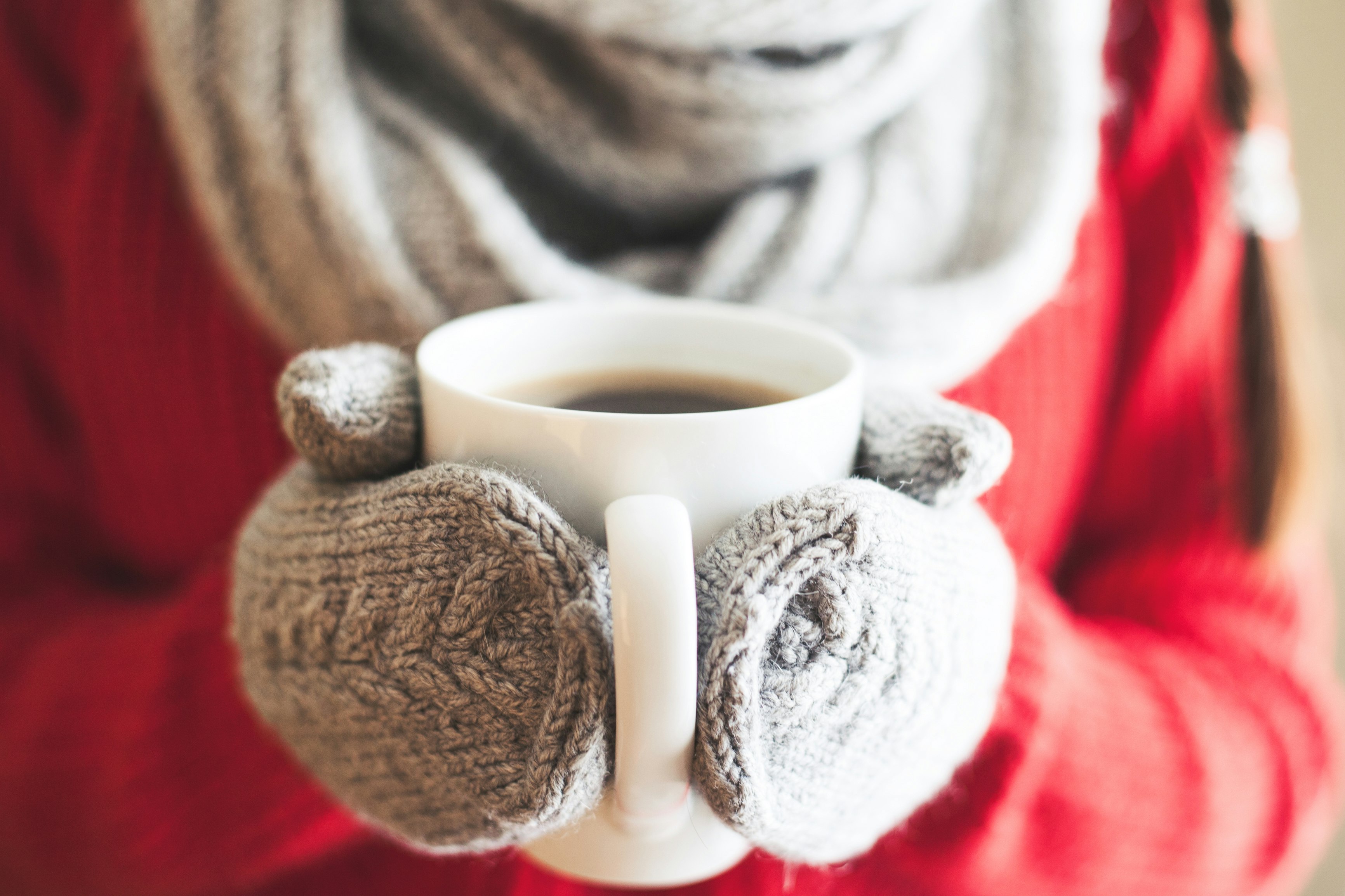 A photograph of a person wearing grey knitted gloves grasping a white mug of drink. In the background the person wears a red jumper and matching grey scarf.