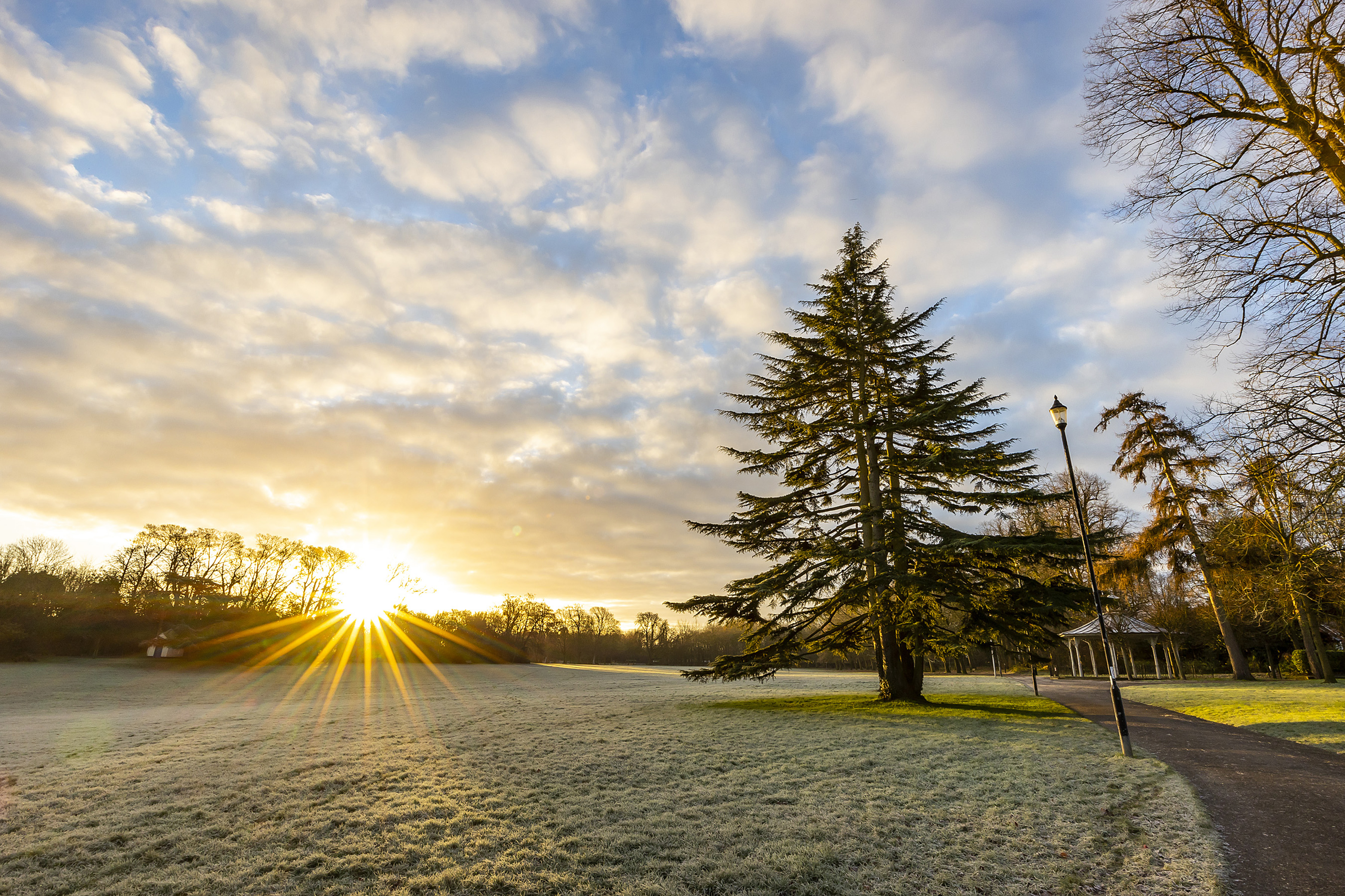 War Memorial Park in Basingstoke pictured in winter. The ground is frosty, the sun is rising behind trees from the left hand side of the image. The bandstand is pictured to the right.