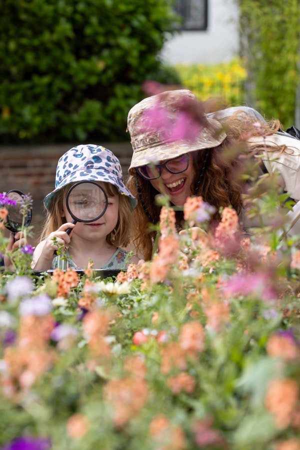 One adult and one child lean into the camera, they are looking through a magnifying glass to find fairies in a bed of flowers.