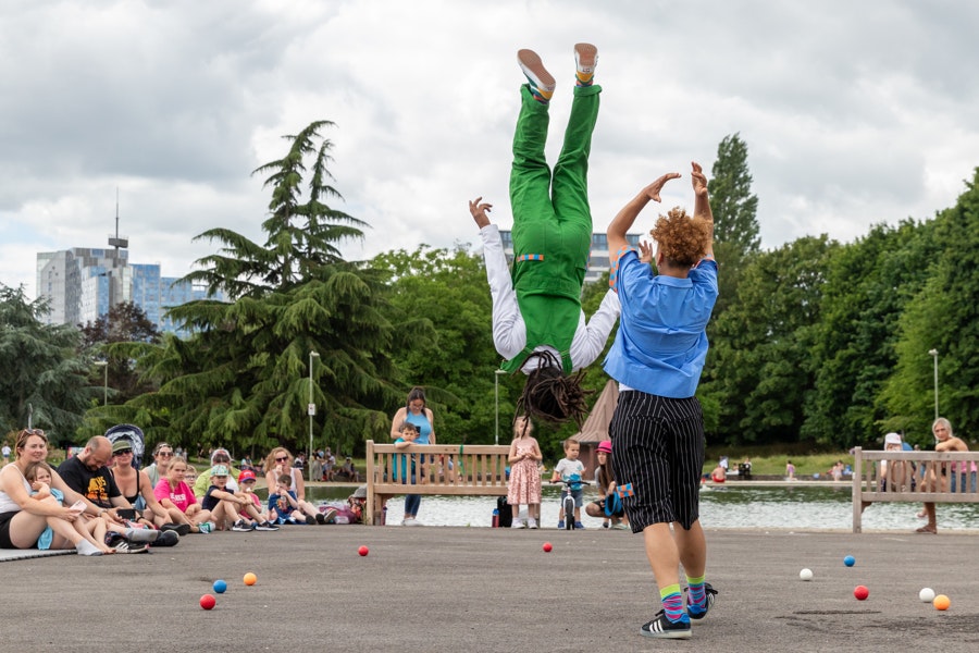 Two people perform outdoors at Eastrop Park in Basingstoke. One wears a blue shirt and flips the other person up in the air. The person in the air wears green dungarees and is upside down facing away from the camera. An audience watch on.