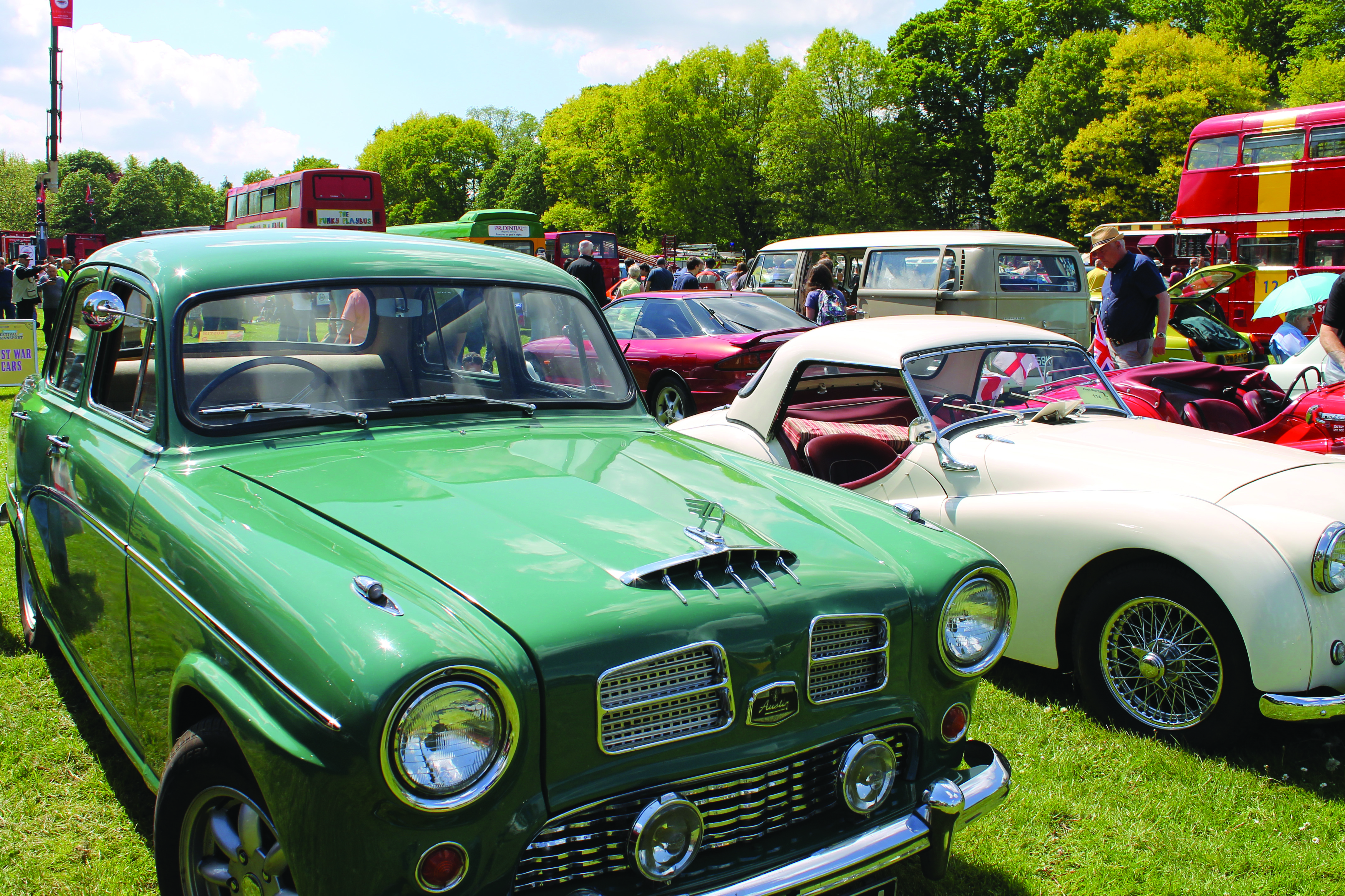 Vintage vehicles are lined up on a field as a display as part of the Festival of Transport. The weather is sunny with white fluffy clouds in the sky.