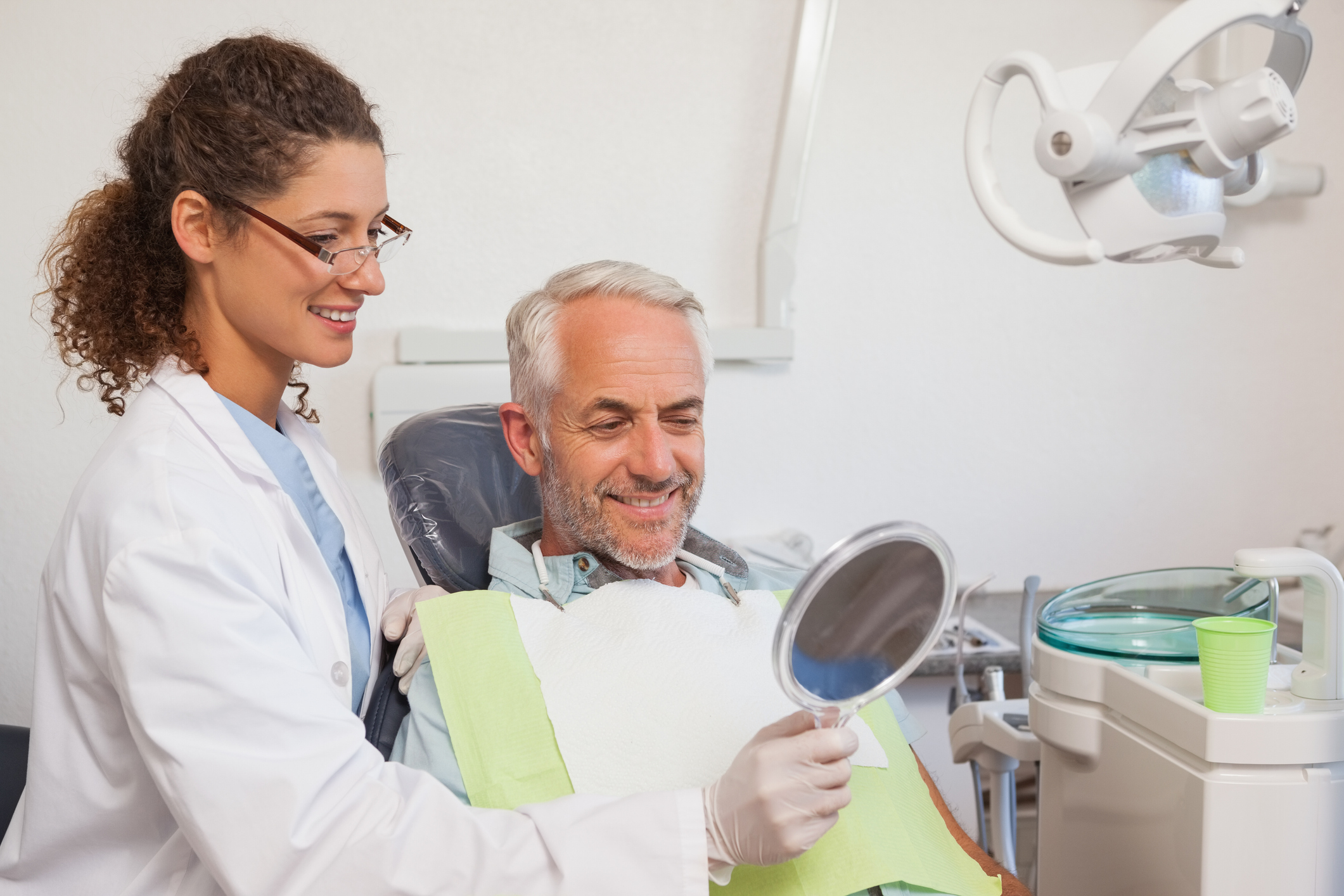 A dentist stands next to a patient, holding a mirror to the patients face. The patient smiles into the mirror. The dentist wears a white gown and glasses.