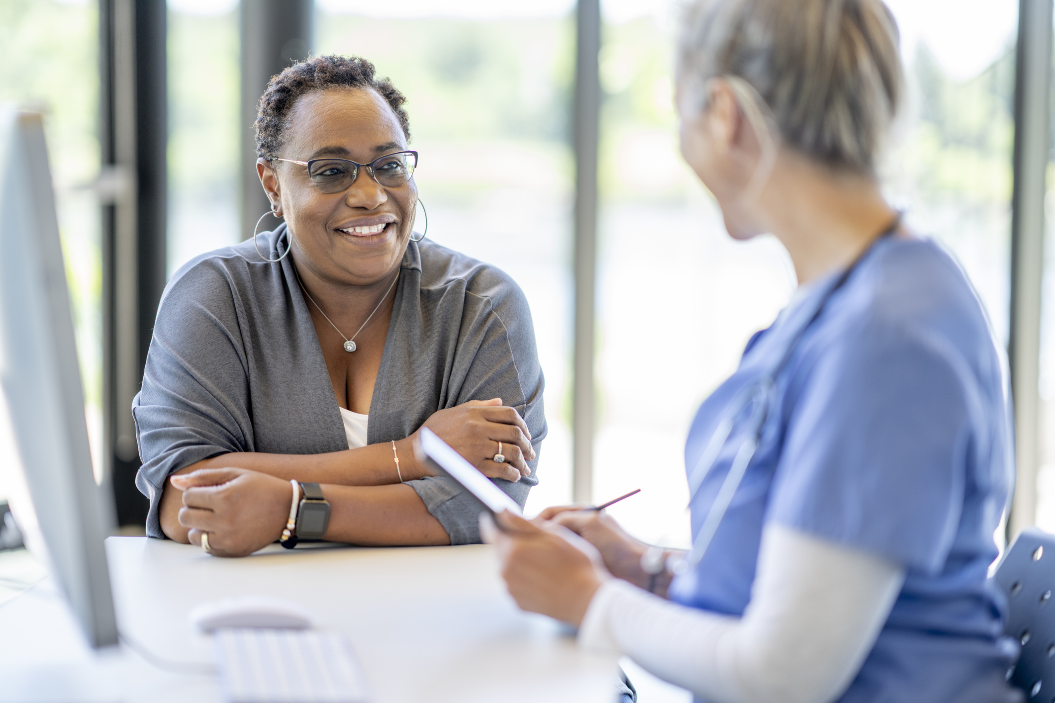 A photograph of a doctor talking to a patient. The doctor has their back to the camera wearing a blue shirt. The patient is face on to the camera and smiles at the doctor.
