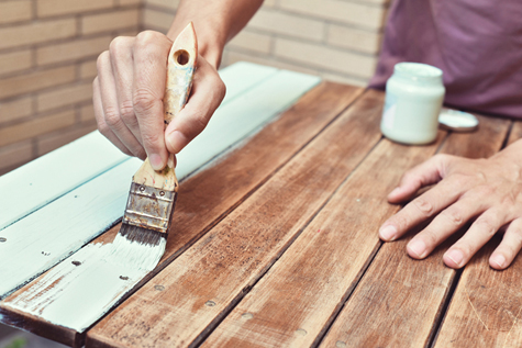 A photograph showing a person upcycling a wooden table. They hold a paintbrush with green paint in one hand. Two of the planks of wood are painted in the new colour duck egg green.