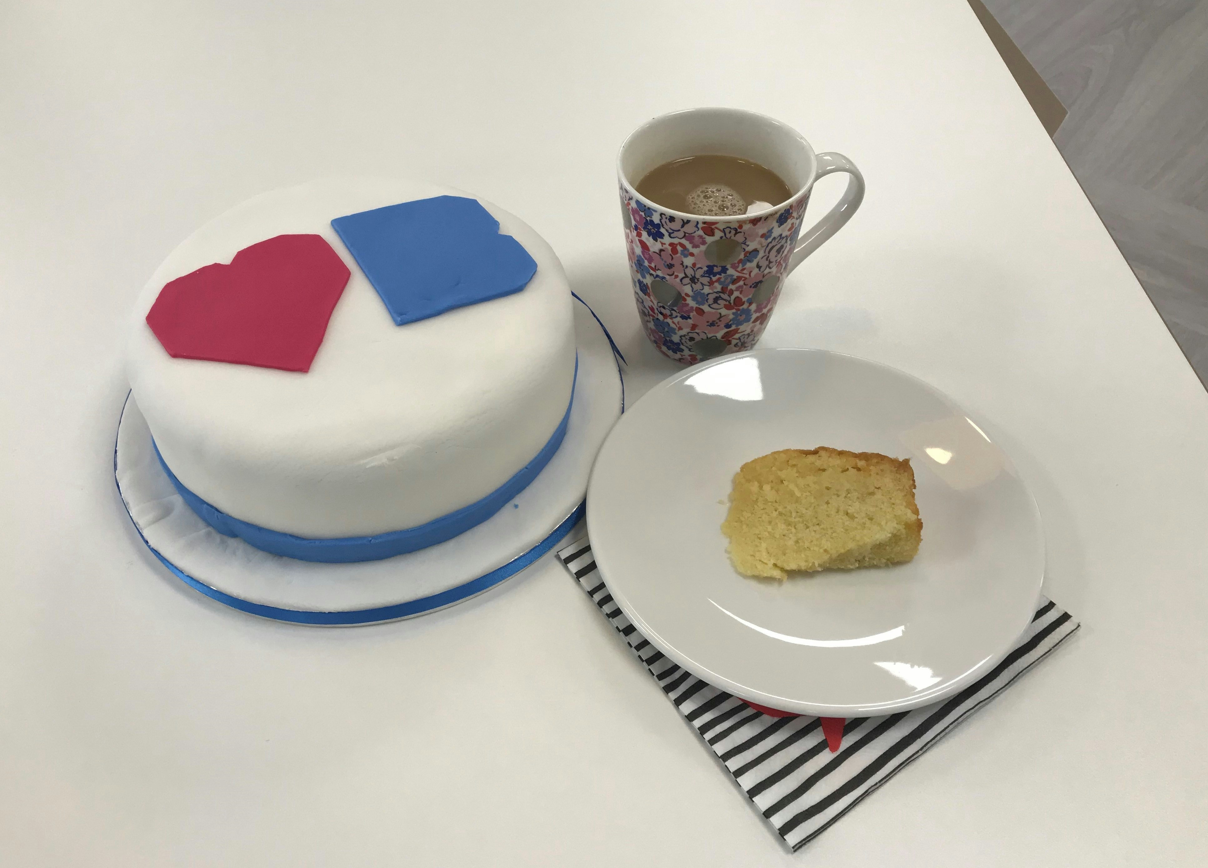 A cake with white icing and the Love Basingstoke logo on. It sits on a cake board with a blue ribbon around the bottom. Next to it is a cup of coffee and another slice of cake that is cut, lying on it's side on a plate. The plate sits on a napkin. All three items sit on a white table.