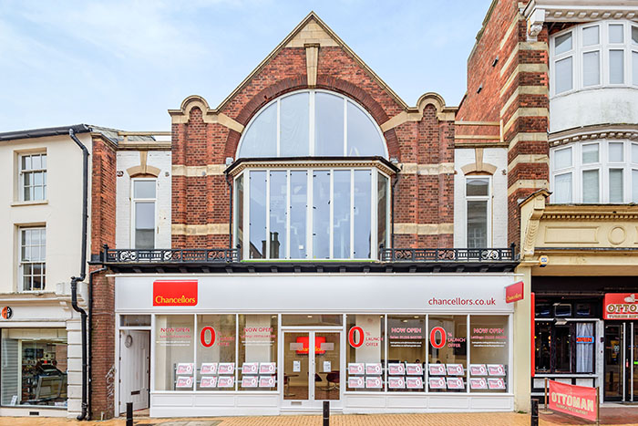 The outside of the Estate agents. A red brick building with large bay window and semi circular window on the top floor. Below at street level is a white and glass front with the Chancellors logo.