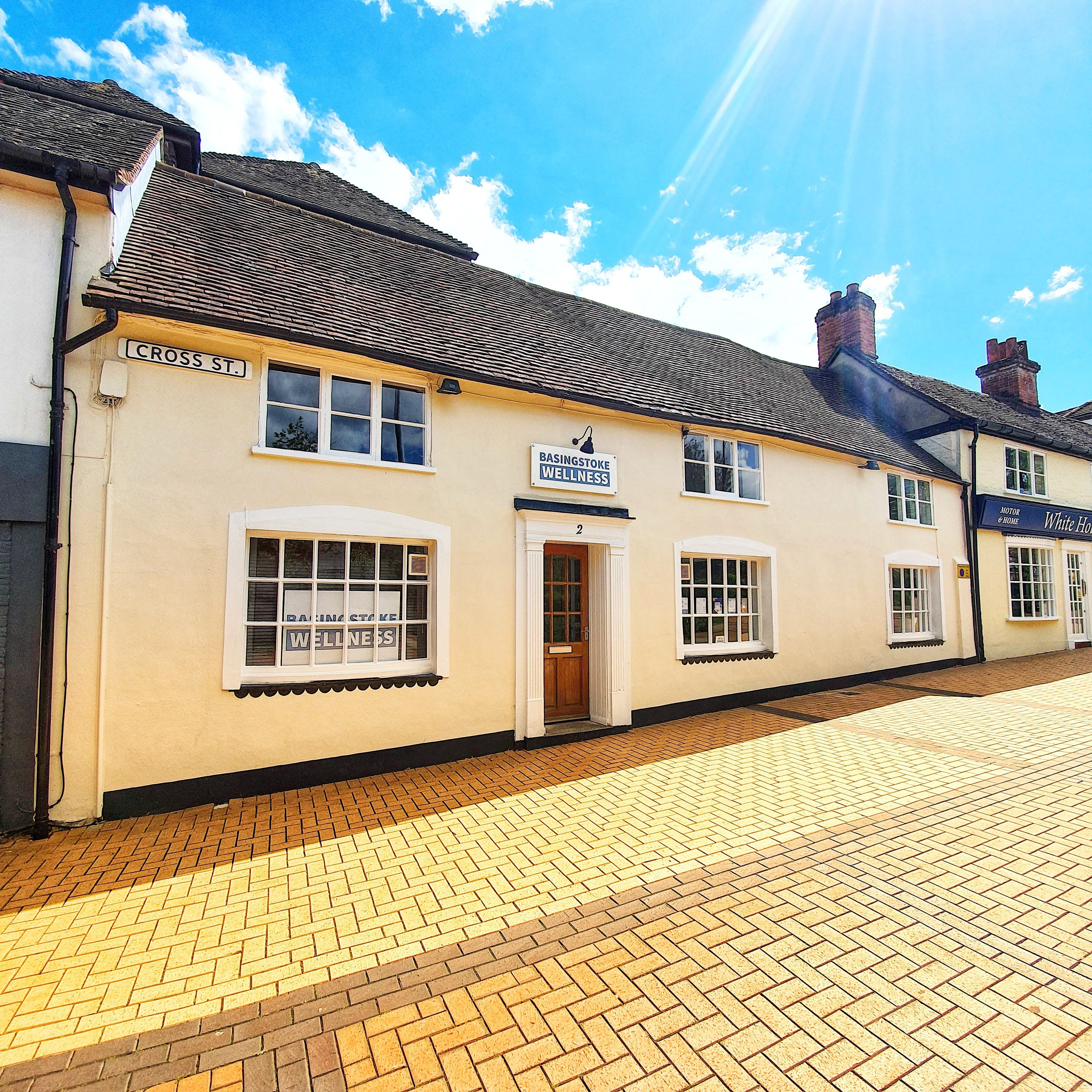 A cream two storey building with white windows and a brown door. The Basingstoke wellness sign is above the door and you can see a street sign for Cross Street on the building. In front of the building is block paving.