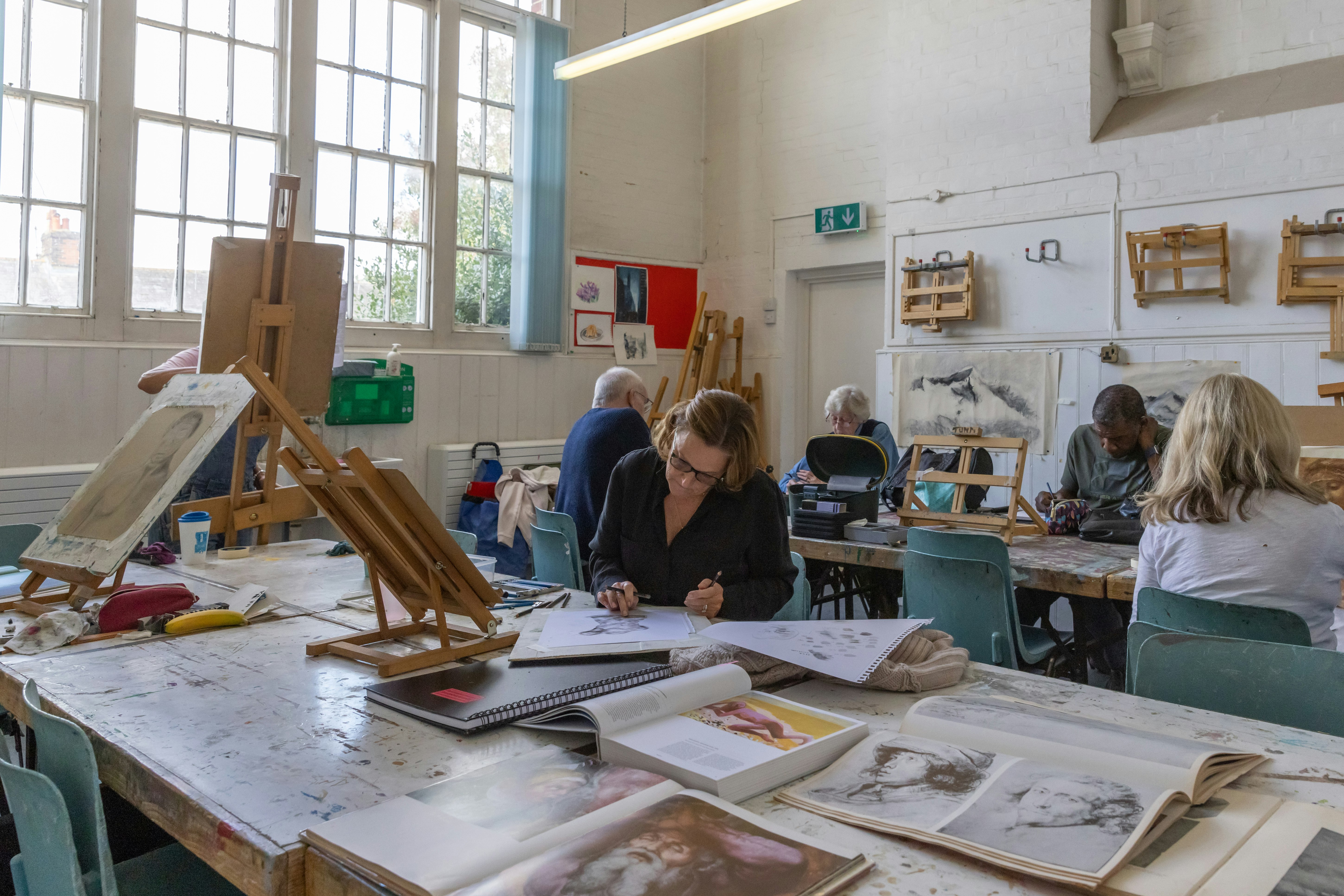 An image of a large well lit room. Five adults can be seen creating art work. They are sat at desks surrounded by paper, art books and easels