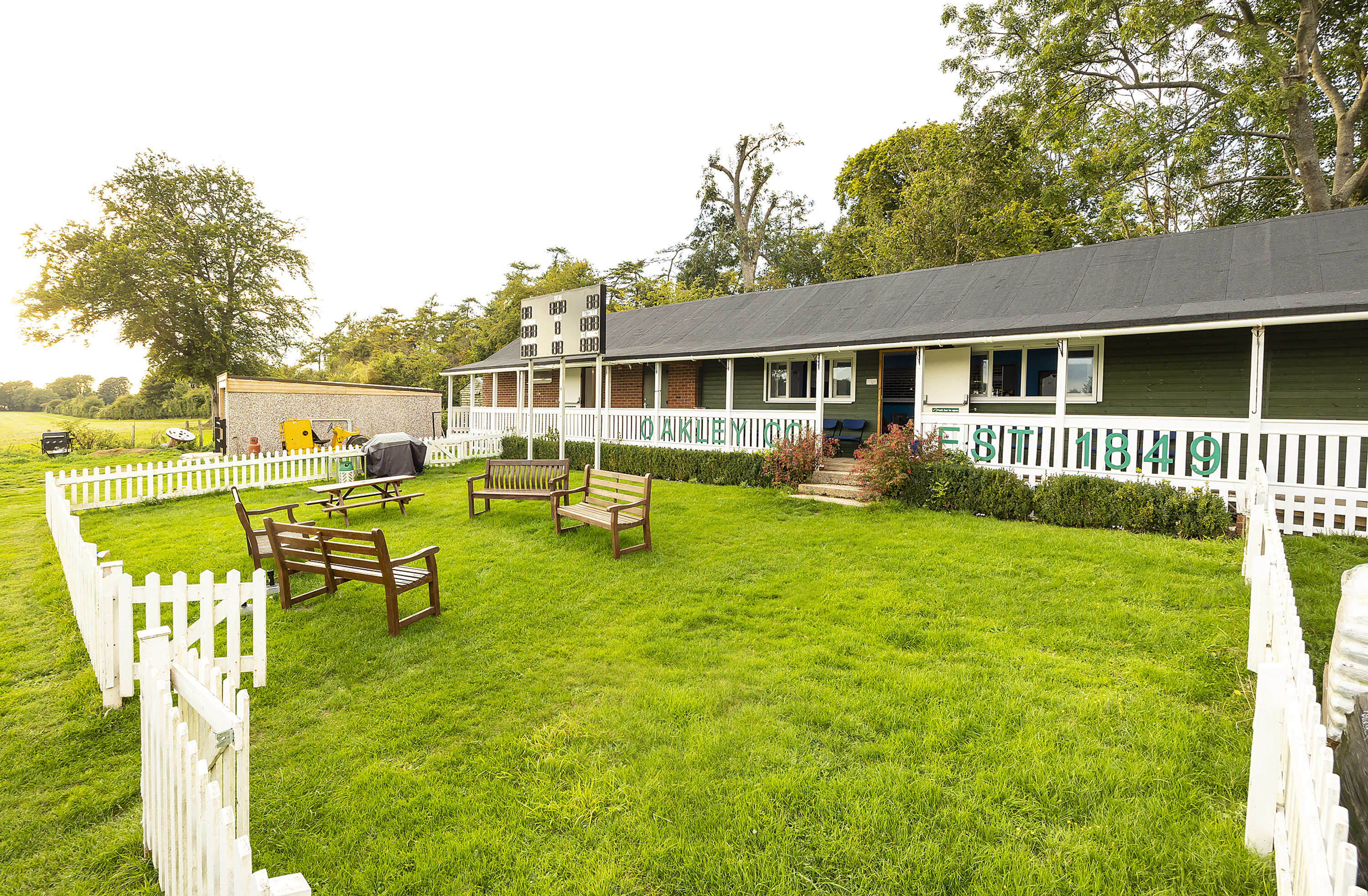 An image of the outside of a white wooden cricket cub. The hut is surrounded by bright green grass and a white wooden fence.
