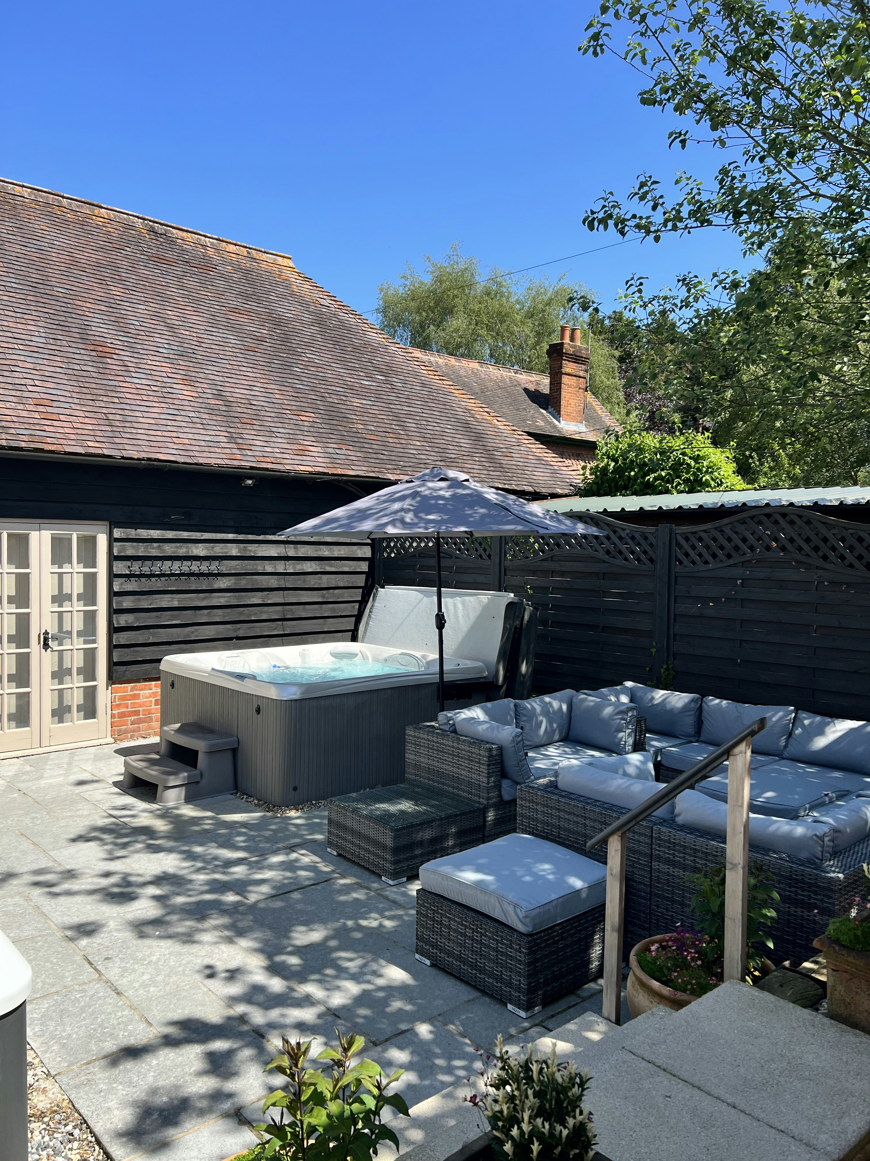 An external image of day spa Laurabella in Basingstoke. The image shows a seating area and hot tub on a paved patio. The sky is blue and in the backdrop is the rear of the building, a tiled roof with wooden cladded walls.