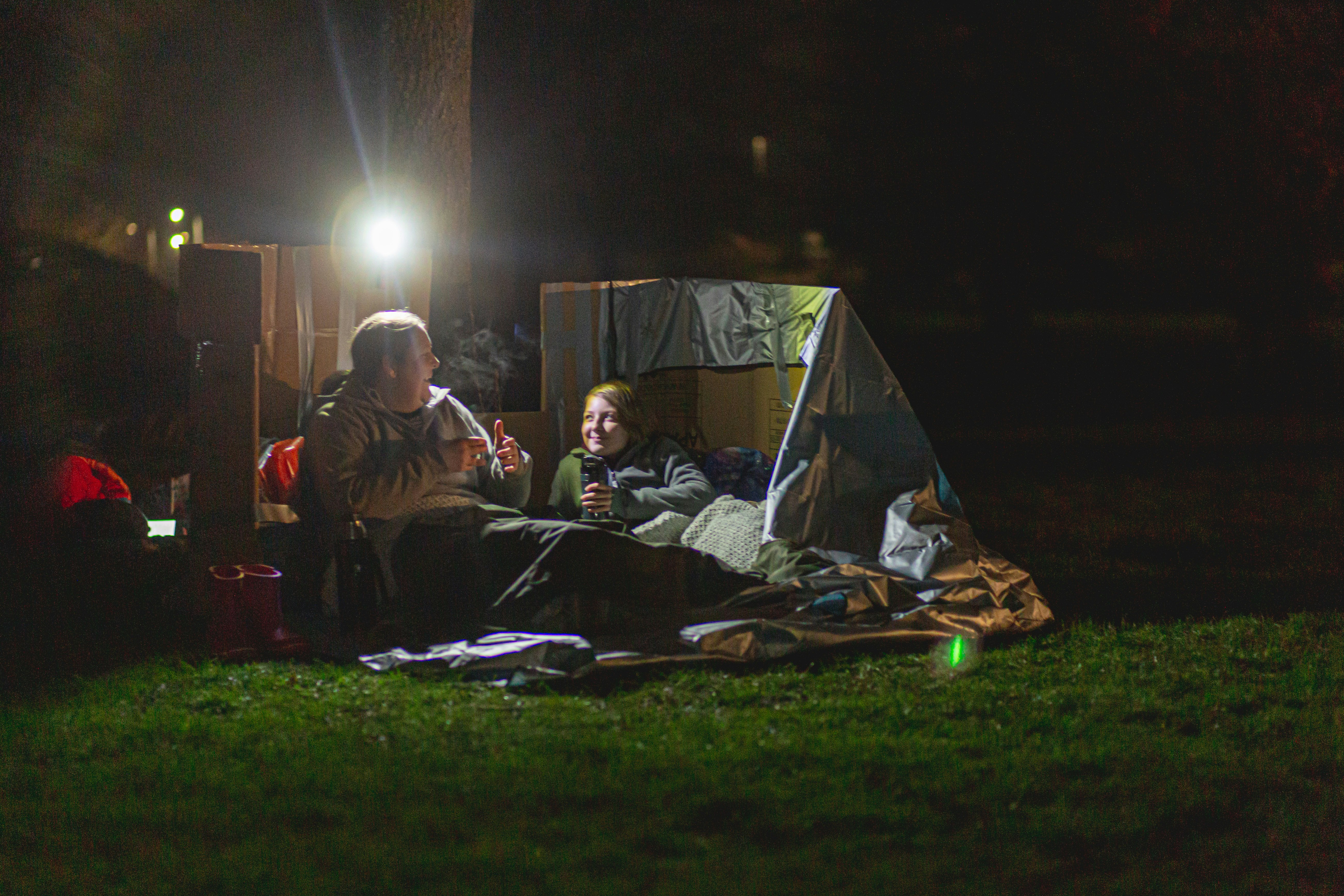 A tent with a man and a child sat together, in the dark outside of a tent. They are huddled in sleeping bags. There is a bright spot light over their heads.