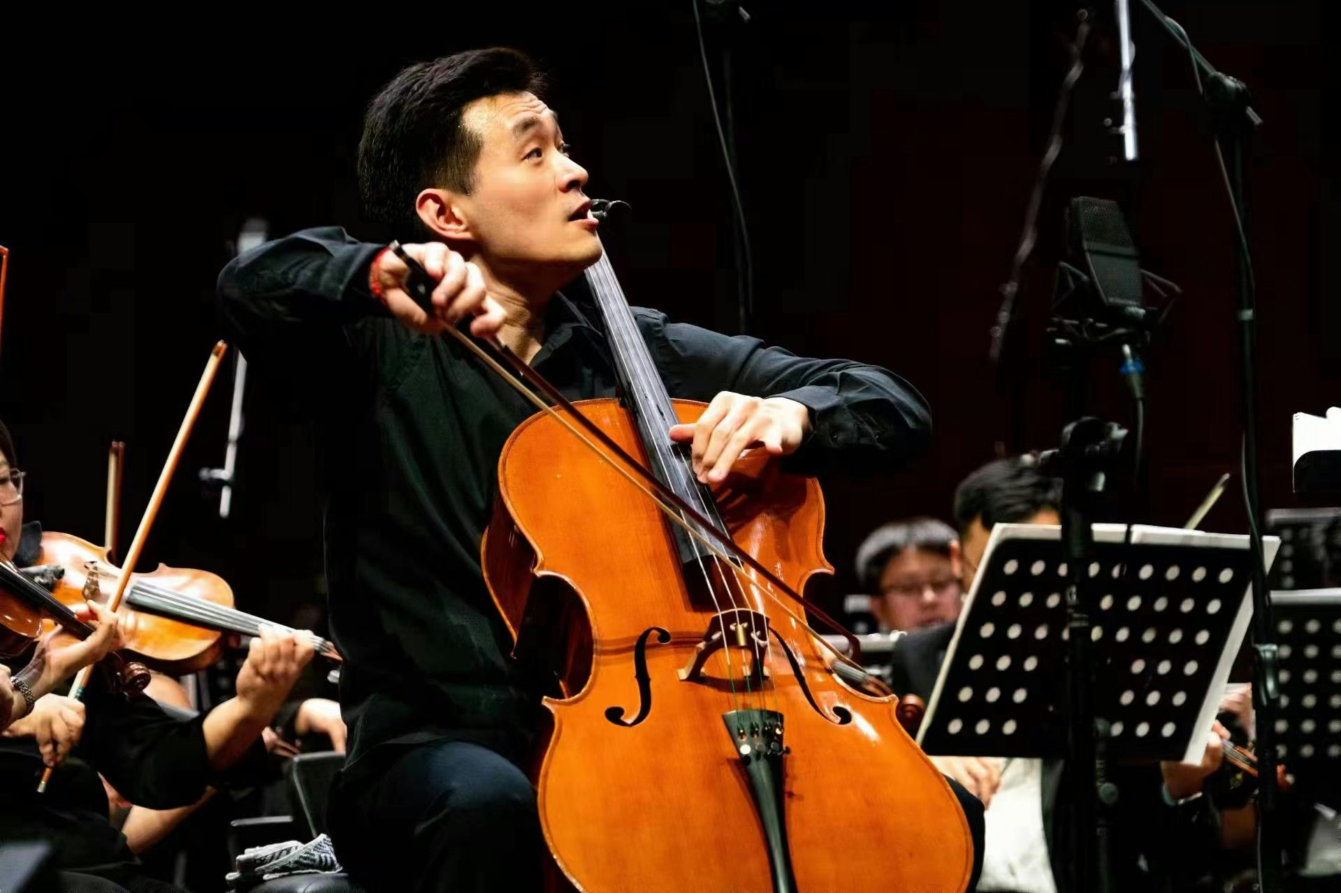 A man with short darrk hair sits on a chair. He is playing a chello in an orchestral performance