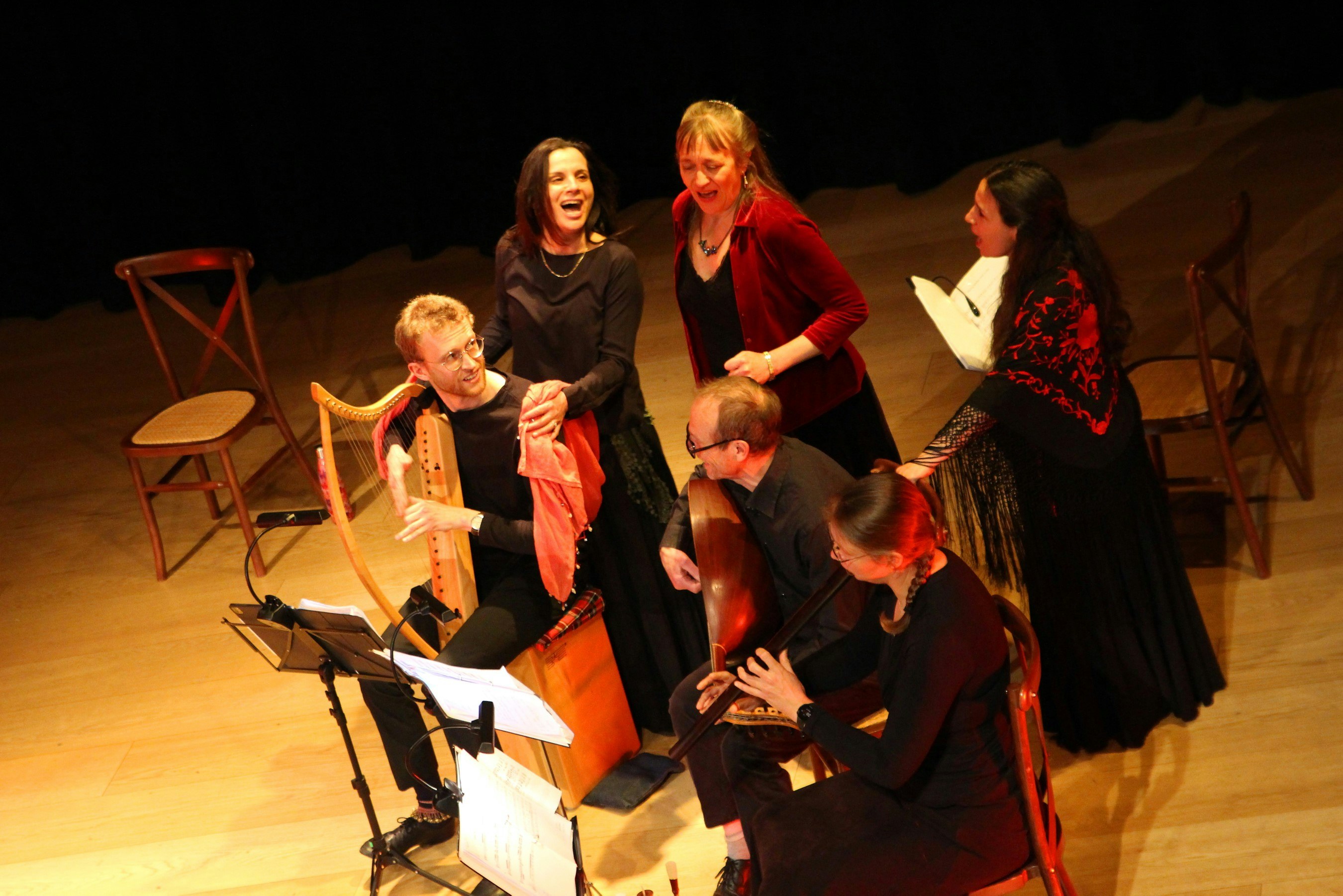 A birds eye view of a stage. On the stage four women and two men are interacting. They are all very animated and two of the people are sat down playing muscial instruments