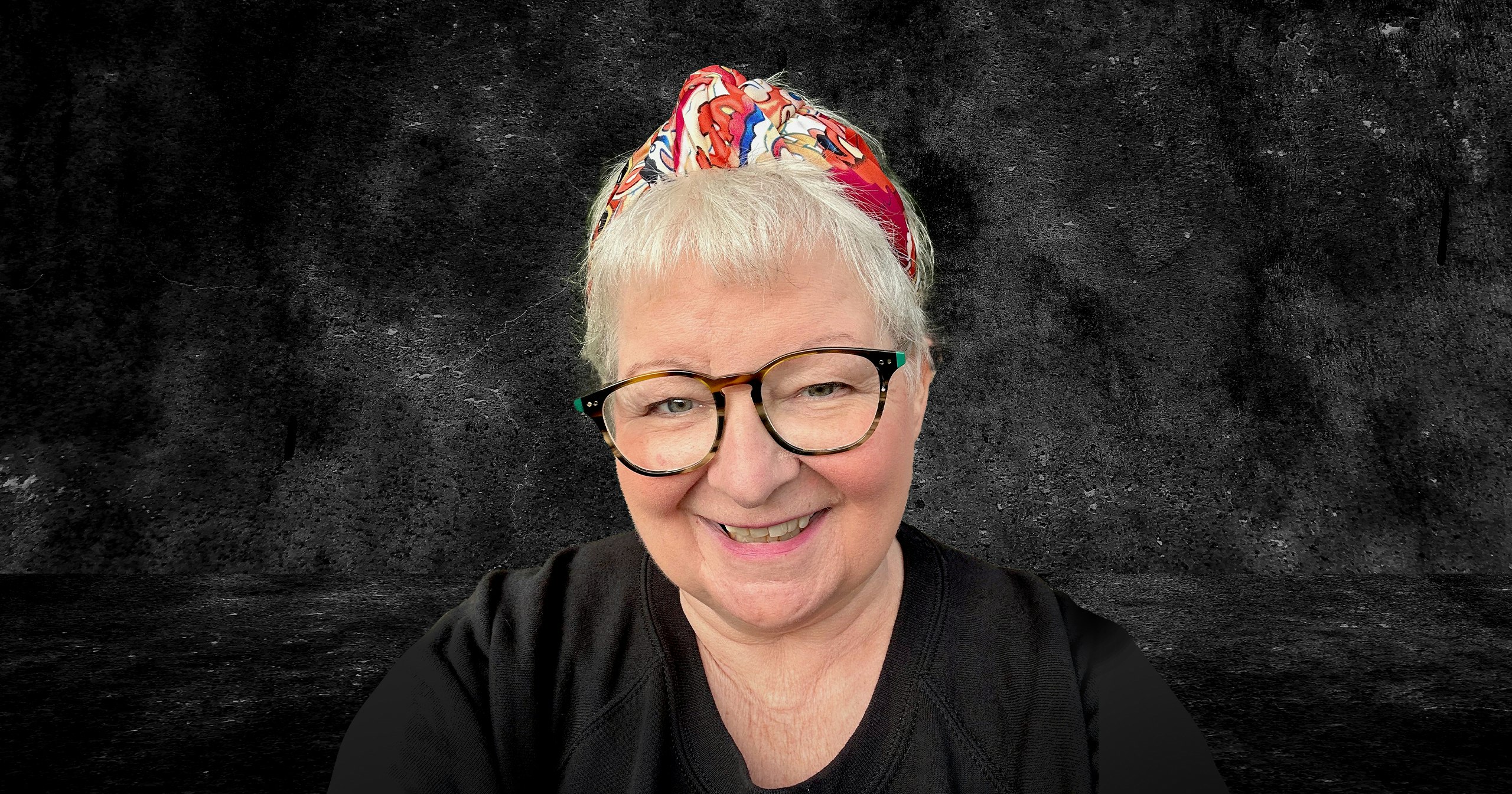 A head and sholders shot of Janey Godley.  She is wearing a black top and glasses with a headband on her short blonde hair.  She is smiling.