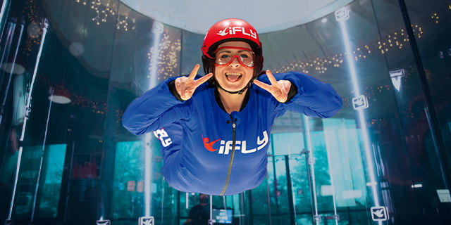 A person smiles at the camera poking their tongue out. They are mid-flight in a chamber at iFLY Basingstoke. They wear an orange helment and blue fly suit.