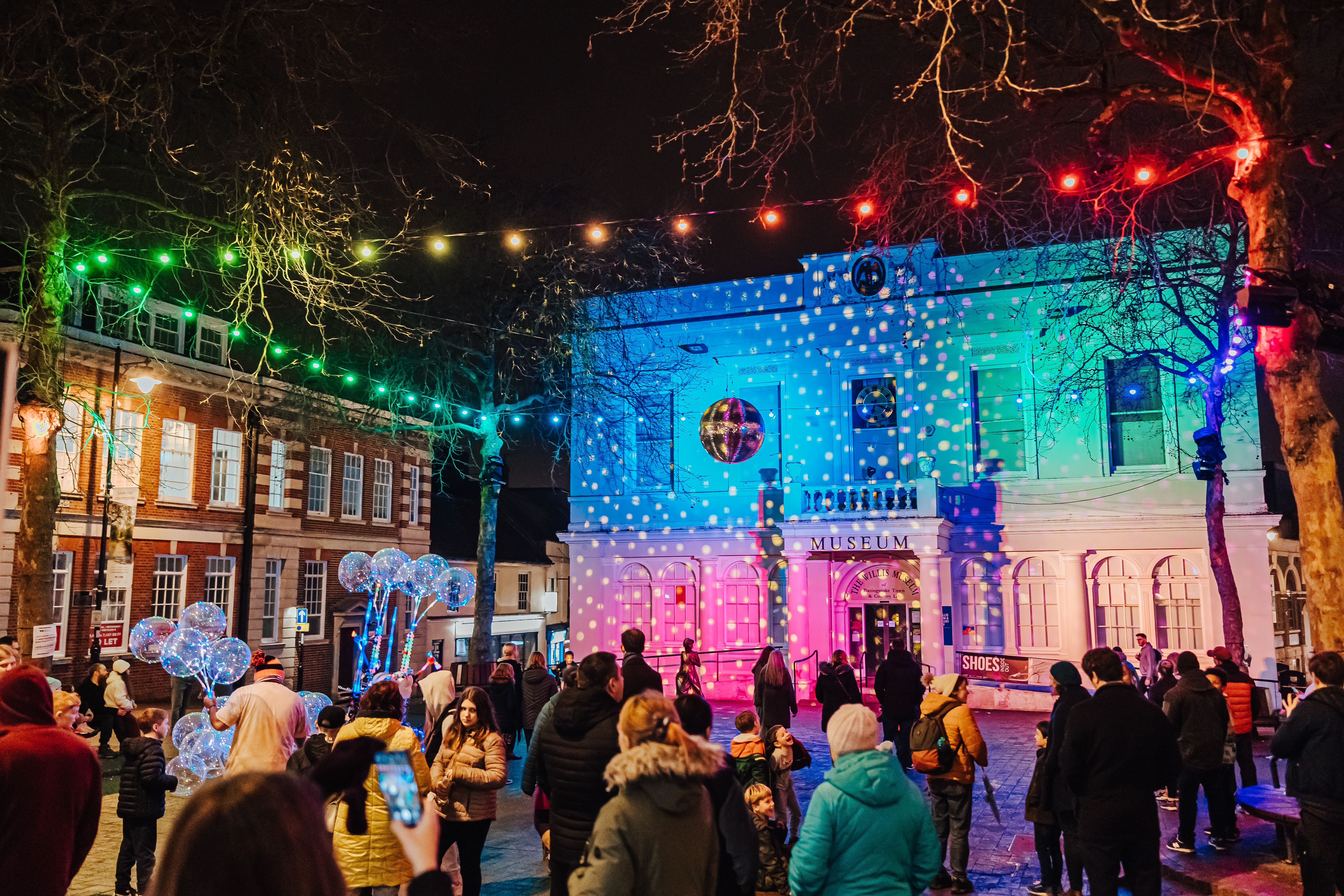 A photograph taken at night. The Willis Museum lit up with a projection. Colourful blue, green and pink lights project onto the front of the museum. Crowds of people stand to the front of the museum. In the air multicoloured festoon lights are strung between the trees. You can see the glitter ball in the middle.