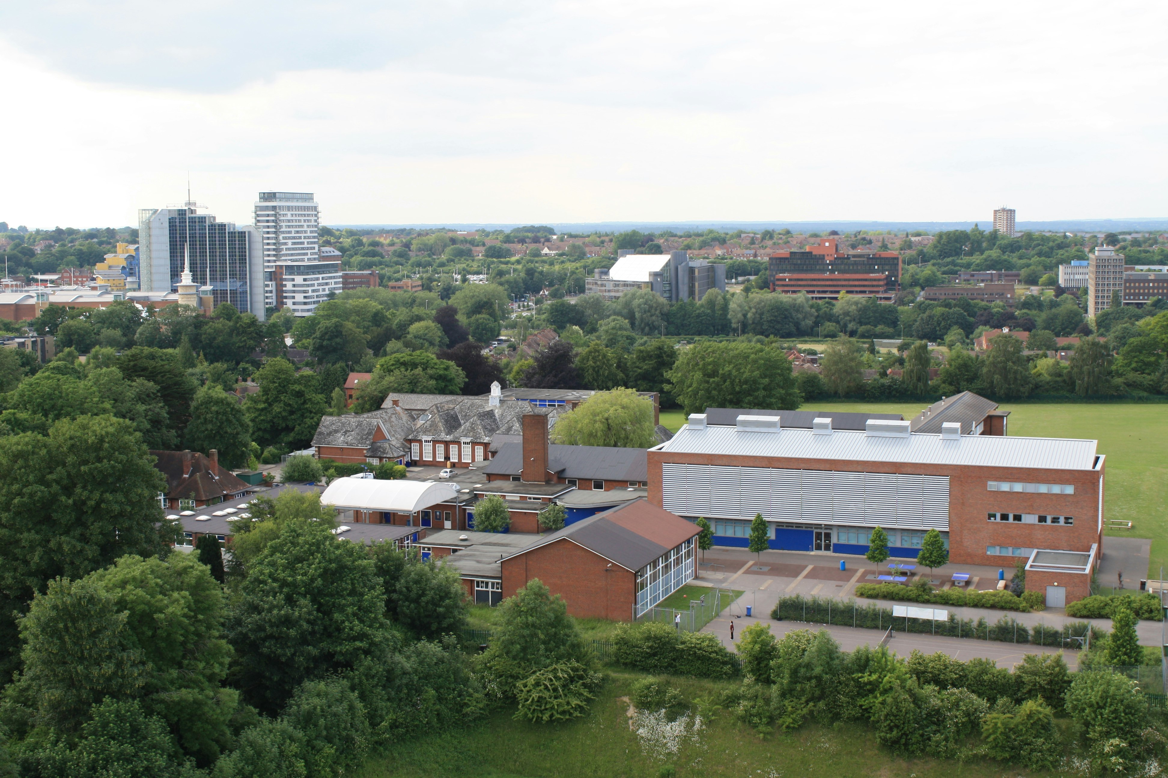 The Costello School from the air. You can see a large building in front with a smaller building on an angle to the left. Behind you can see green fields and in the distance the high rise buildings of Basingstoke town centre.