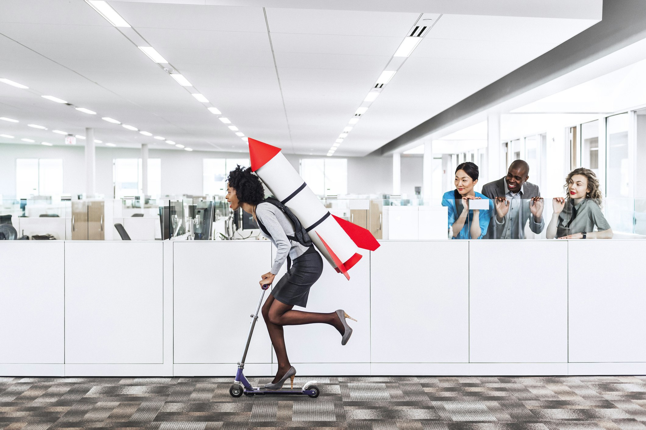 A female is on a scooter in an office and has a rocket on her back. Three people look on.