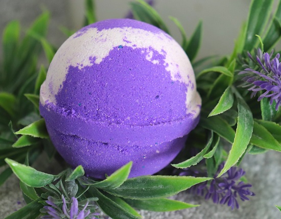 A purple and white bathbomb sitting on a bed of green leaves and purple flowers.