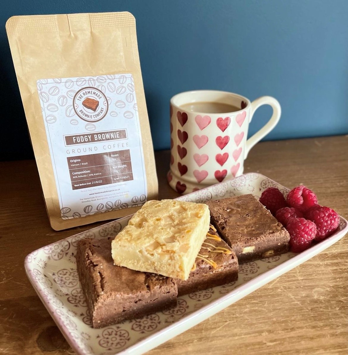 A tray of brownies with raspberries, a mug and a pouch of coffee.