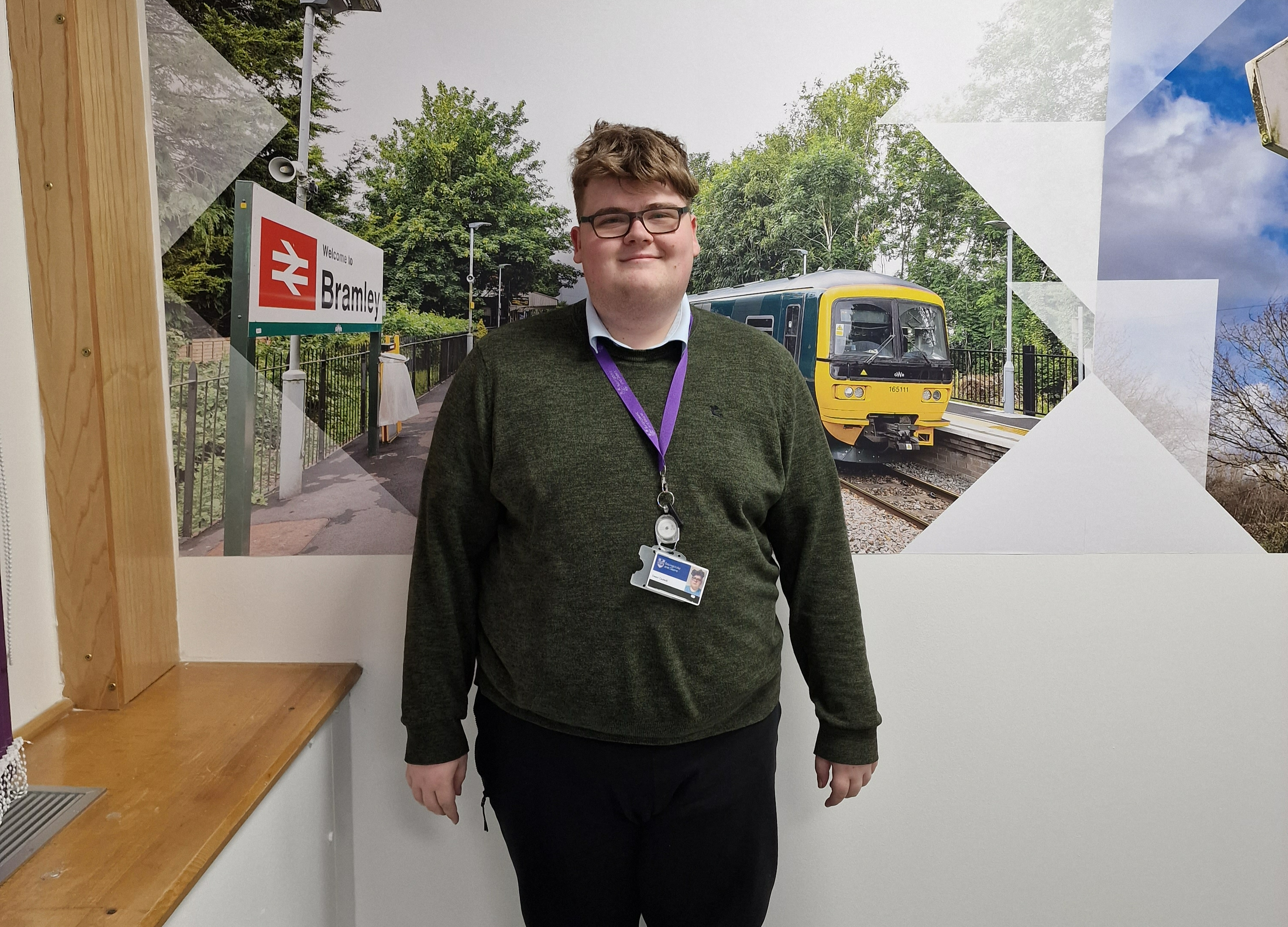 A person stands in front of some photos. He wears a green jumper.