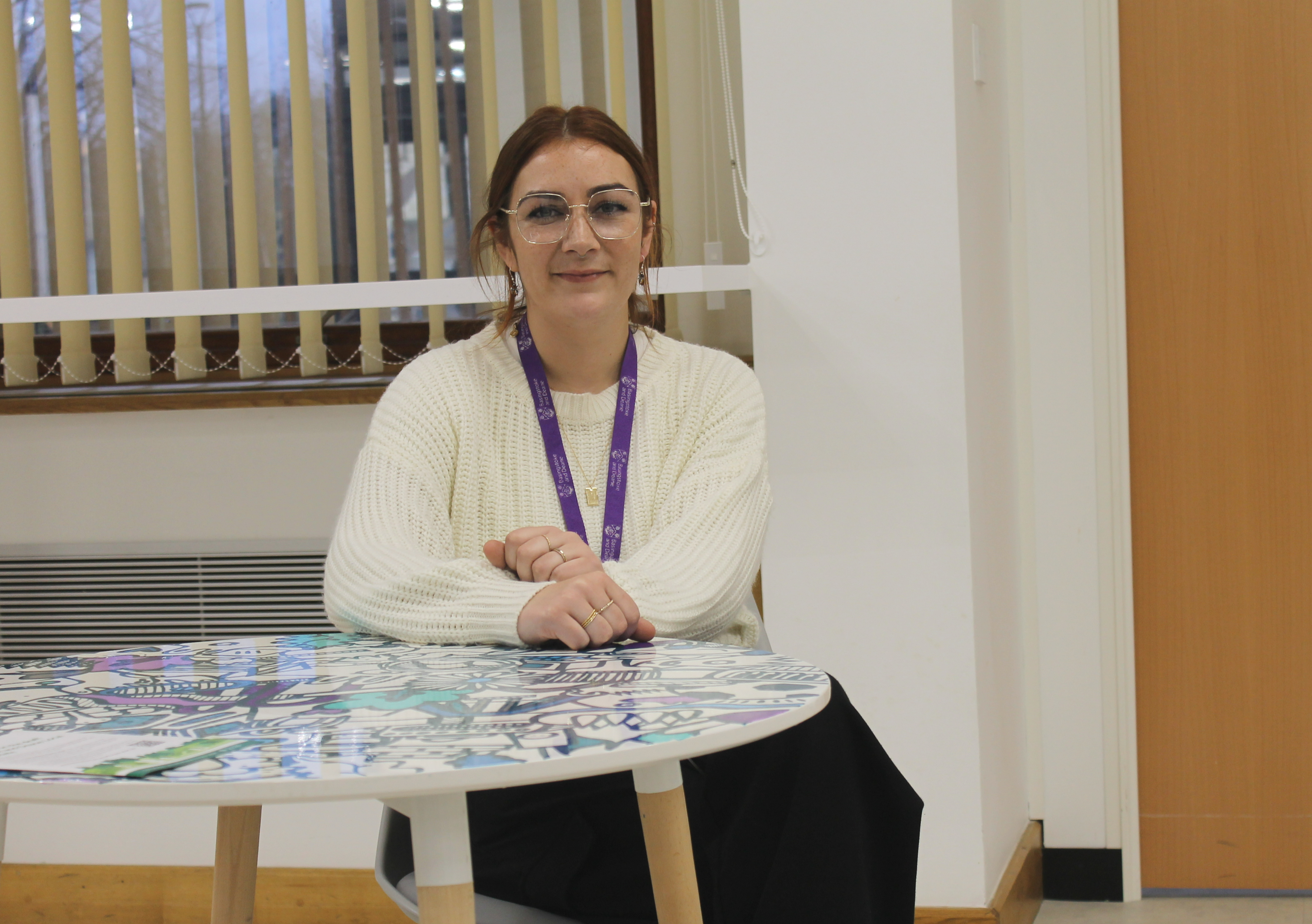 A female sits behind a table with her hands resting on it. She wears a white jumper.