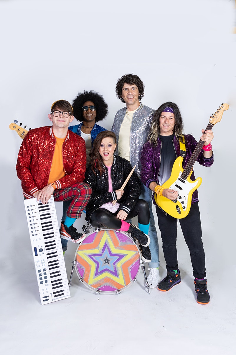 there are five people stood together in a huddle. They are all facing the camera and smiling . One person is holding a yellow electric guitar and another is holding a keyboard, whilst the lady in the middle in sat on a drum set.