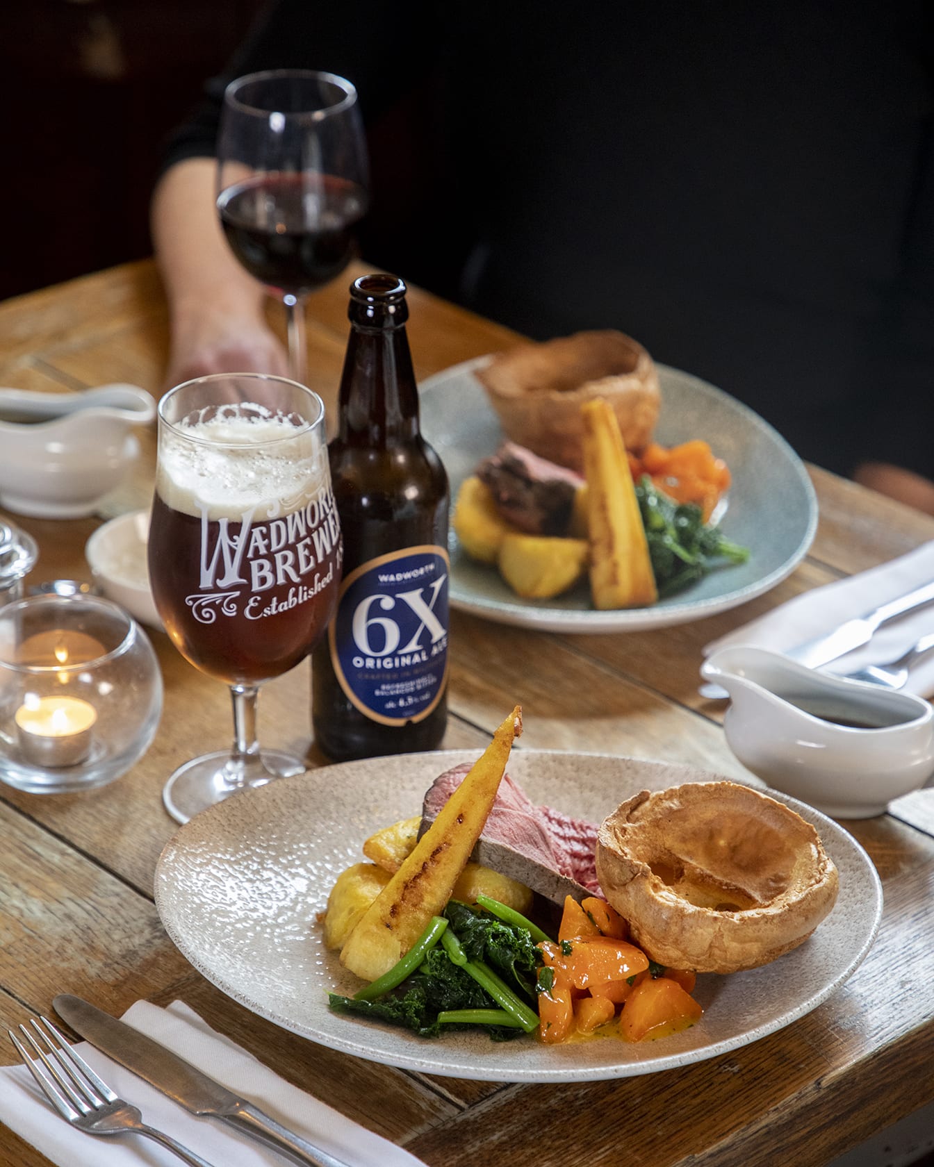 A photograph taken inside Bartons Mill. The image shows two roast dinners set on a wooden table. To the top right of the image is a hand holding a glass of red wine. There is also a bottle and glass of beer on the table next to one of the plates.