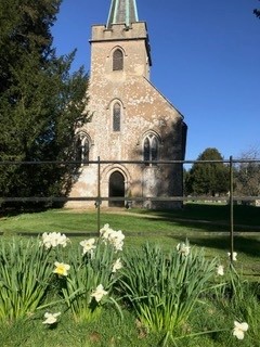 A photograph taken on a sunny day, in the backdrop the historic Steventon church is pictured. Five windows are visible and an arched wooden door. Green grass with pretty daffodils line the front of the church.