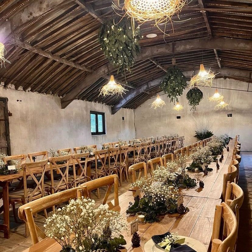 A photograph of a room ready for a wedding reception. The Loft style room has rustic plain walls, a dramatic wooden curved roof and straw lampshades. Two long wooden tables and wooden chairs run the length of the room. The table decorations are simple with white flowers placed in the centre of the tables.