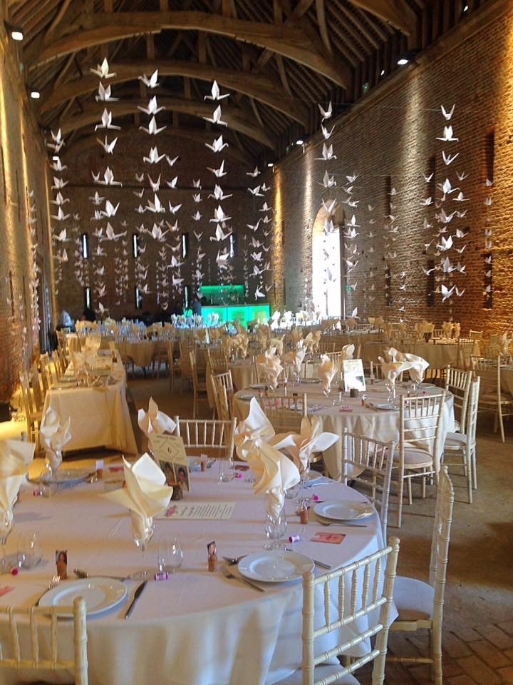 A photograph of Basing House decorated for a wedding. Several round tables, covered in white tablecloths fill the room. White wooden chairs surround the tables. Each table is laid with a white plate, cutlery, cream napkin and a printed number placed in the centre of the table. White origami birds hang from the wooden beamed ceiling.