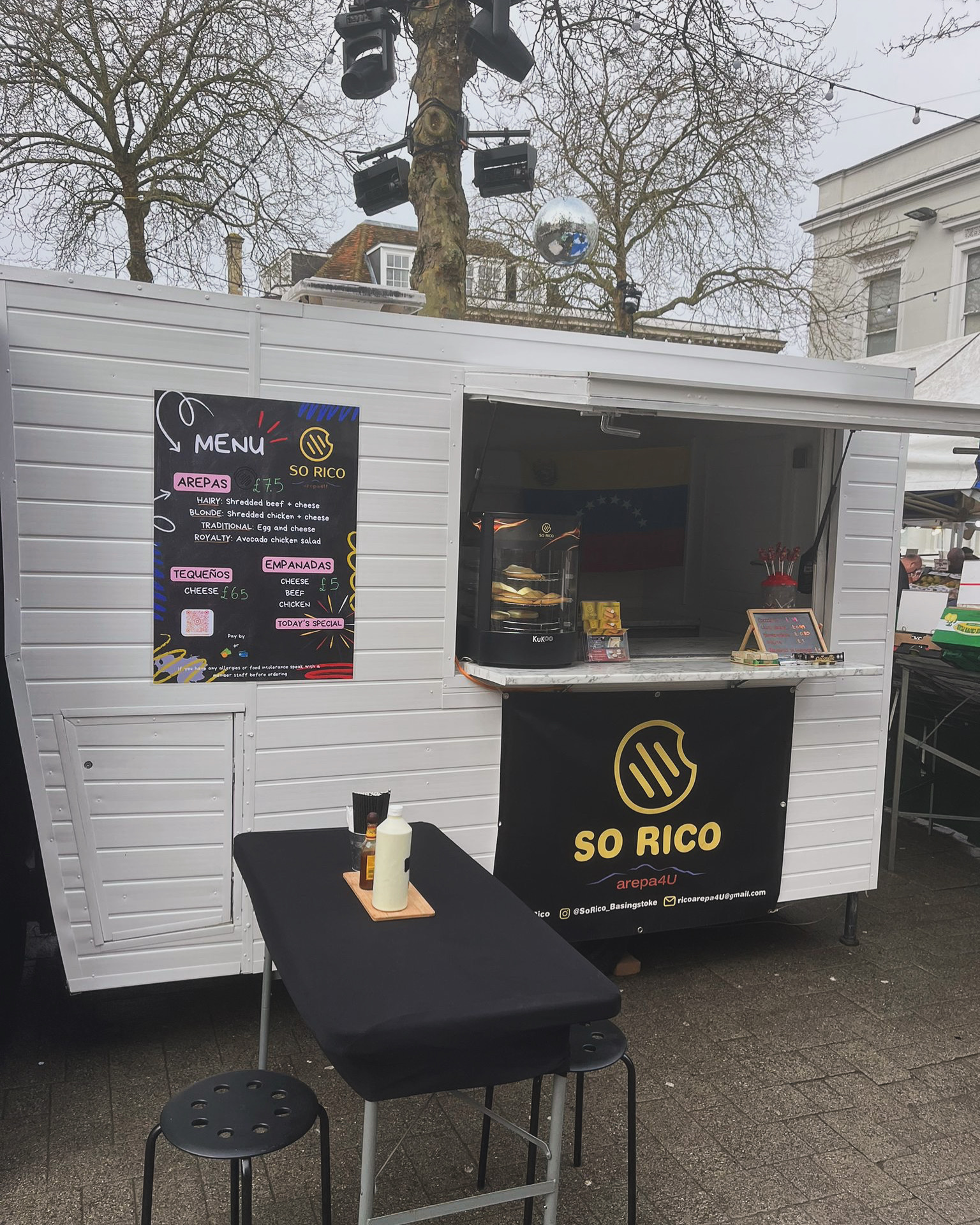 A mobile food van that is white in colour with black signage and an open window. Outside is a black table and stools.