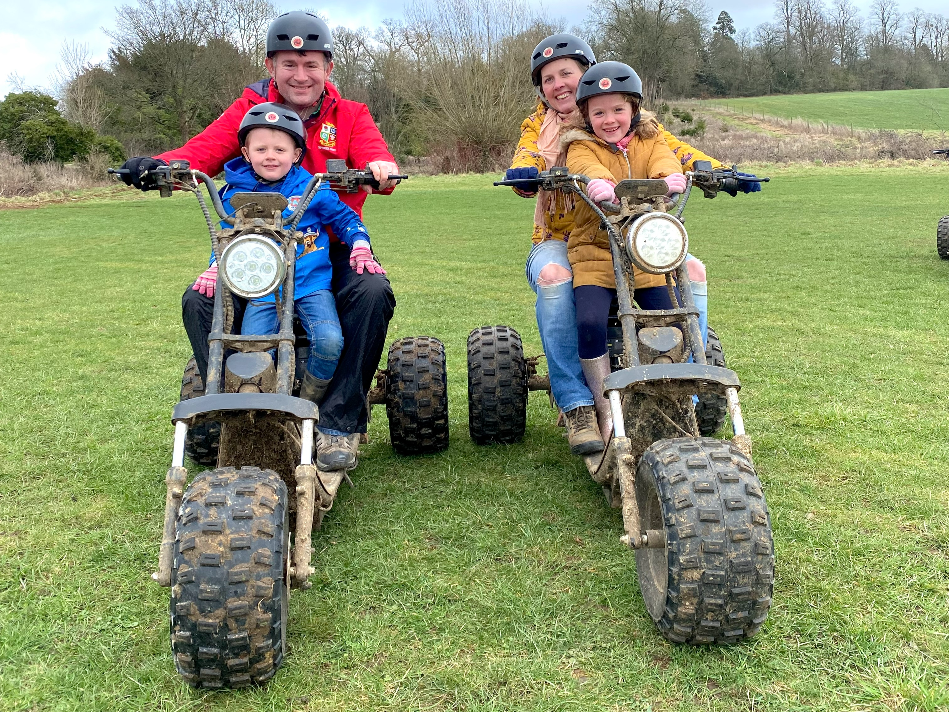 Two adults sit on three wheeled scooters with children in front of them.