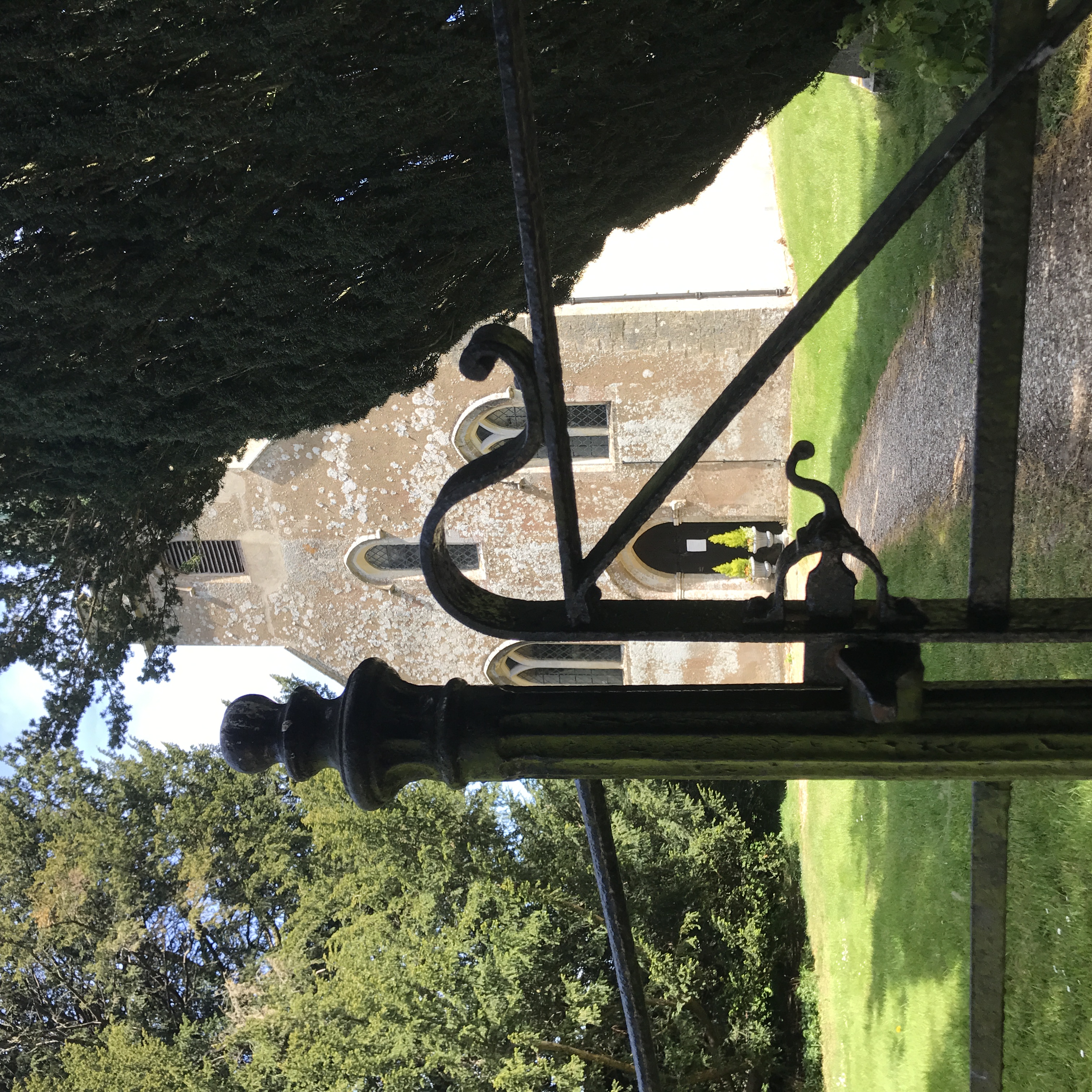 A photograph of Steventon Church. To the front of the image is a set of black iron gates, behind the gates Steventon Church is visible. There are two green trees either side of the church.