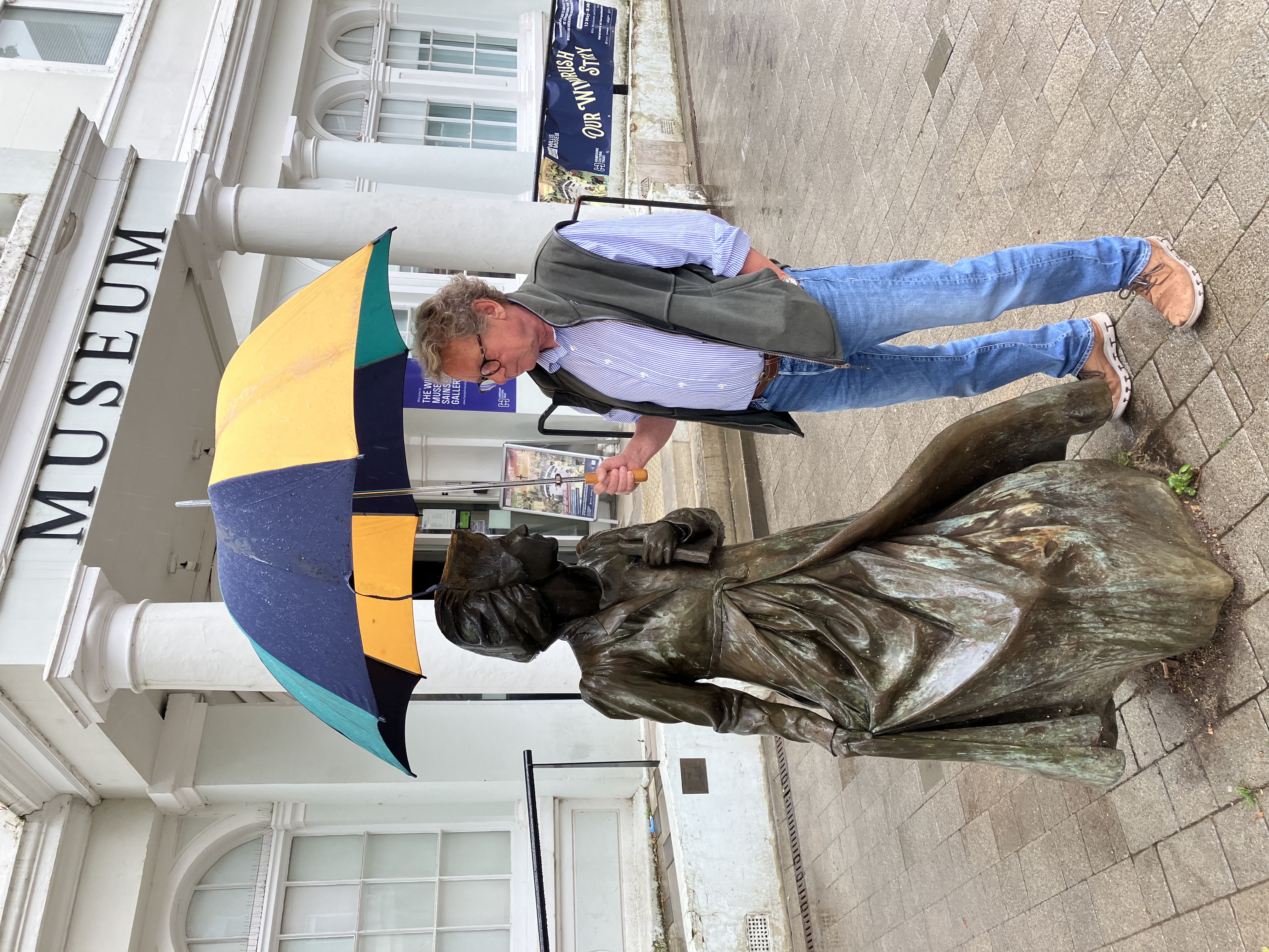 Phil Howe, stands next to the bronze statue of Jane Austen. He holds a colourful umbrella over his head as it is a rainy day,