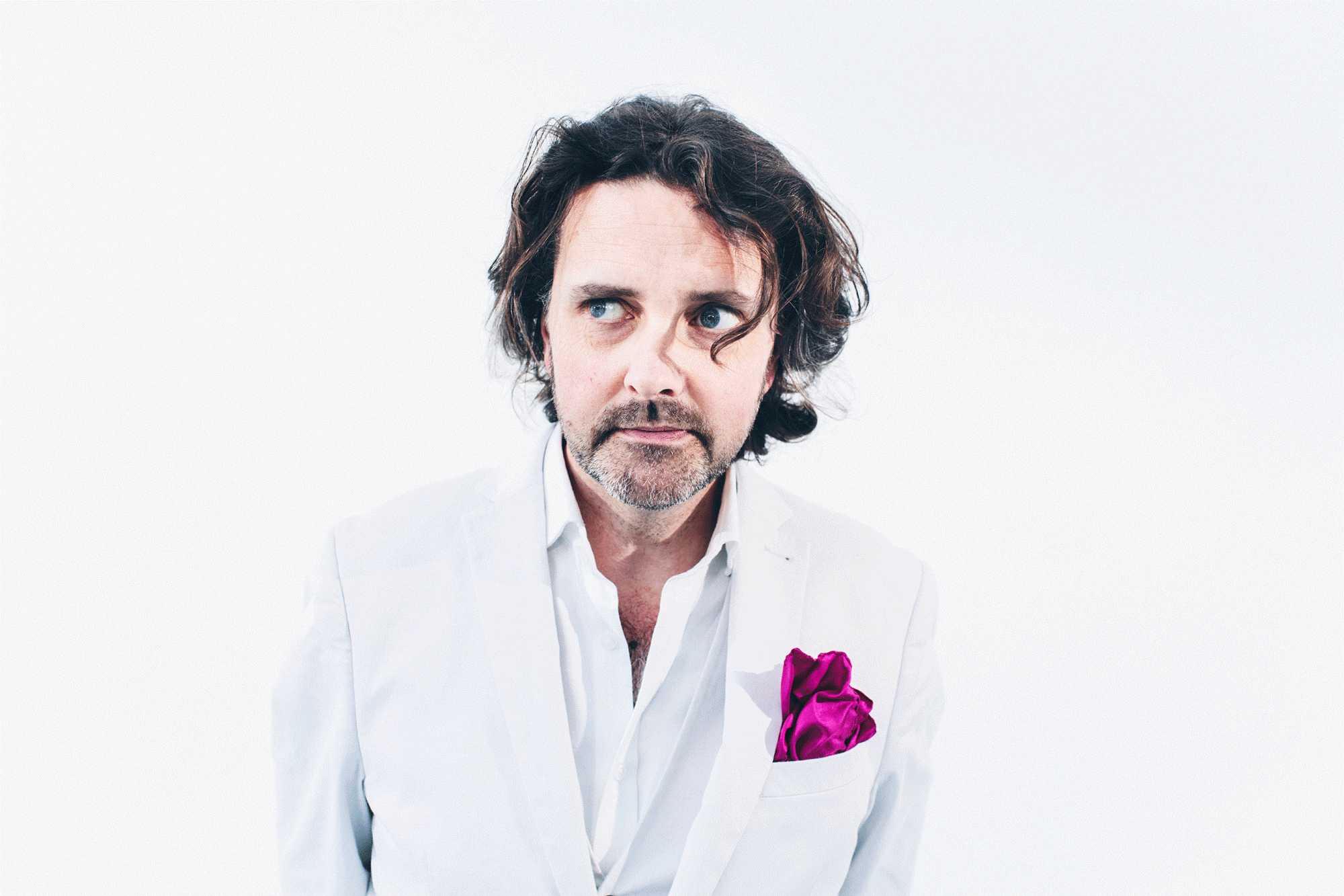 A male wears a white shirt and white jacket with a pink scarf in his pocket. He looks away from the camera. The background is white.
