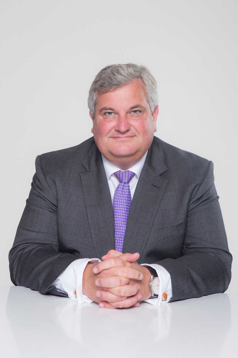 A male wears a suit and looks at the camera with arms on the table.