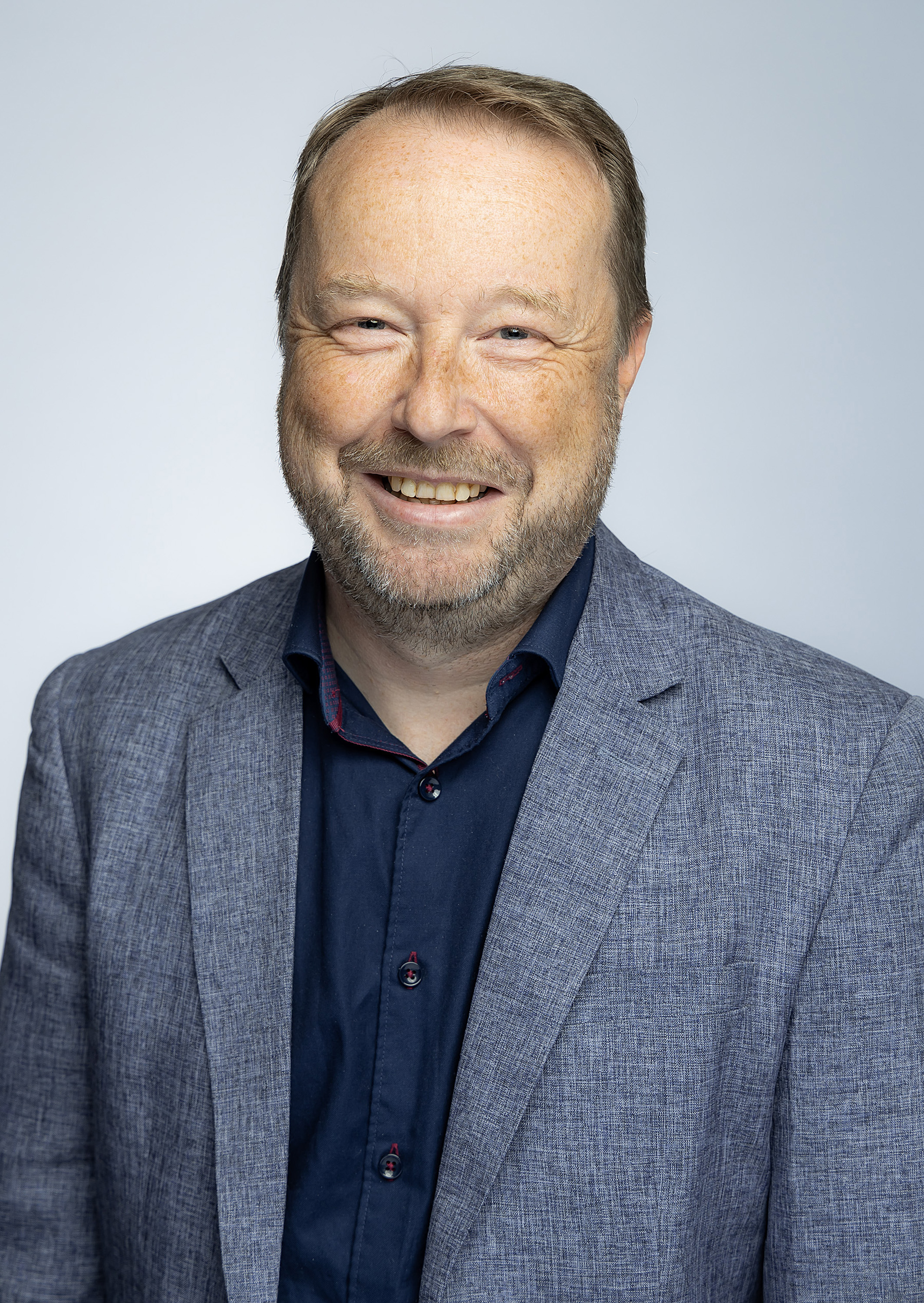 A male wears a grey shirt and jacket and looks at the camera smiling.