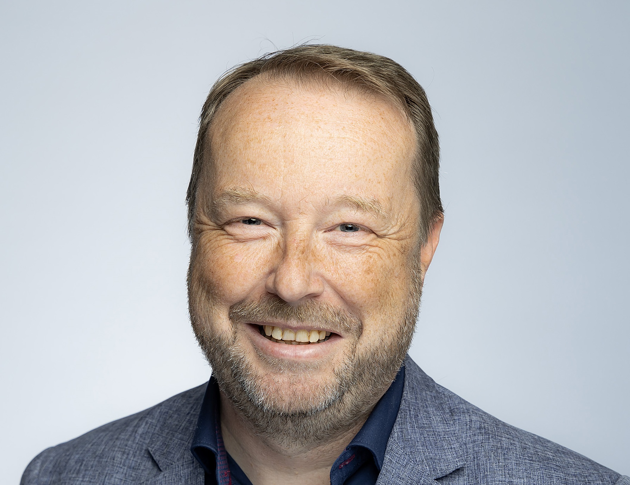 A man wears a blue shirt and grey jacket and looks at the camera.