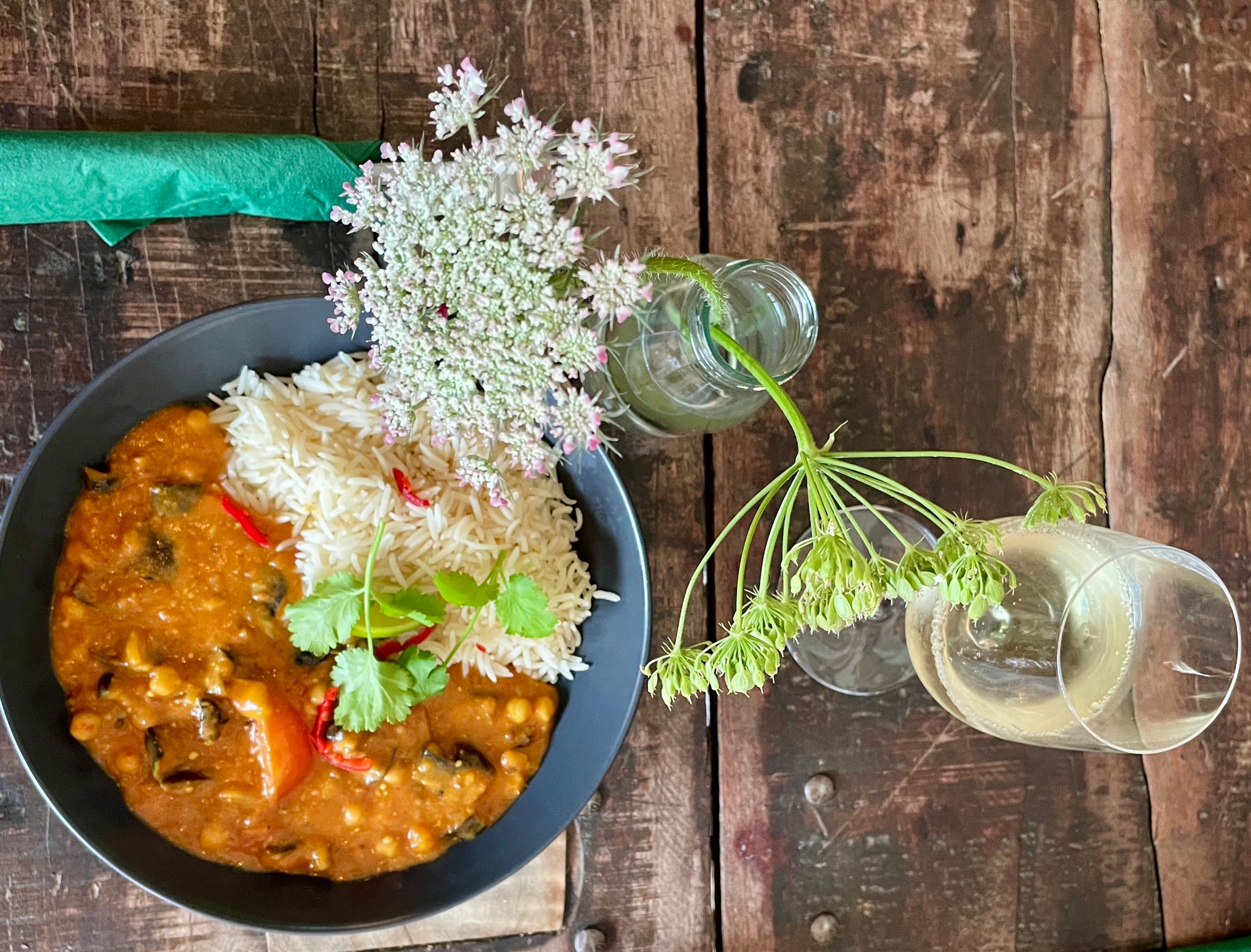 Photograph of a table of food. A bowl full of vegetable curry and rice is laid upon a wooden table. A glass of white wine and a small vase with a wild flower is infront of the bowl of food.