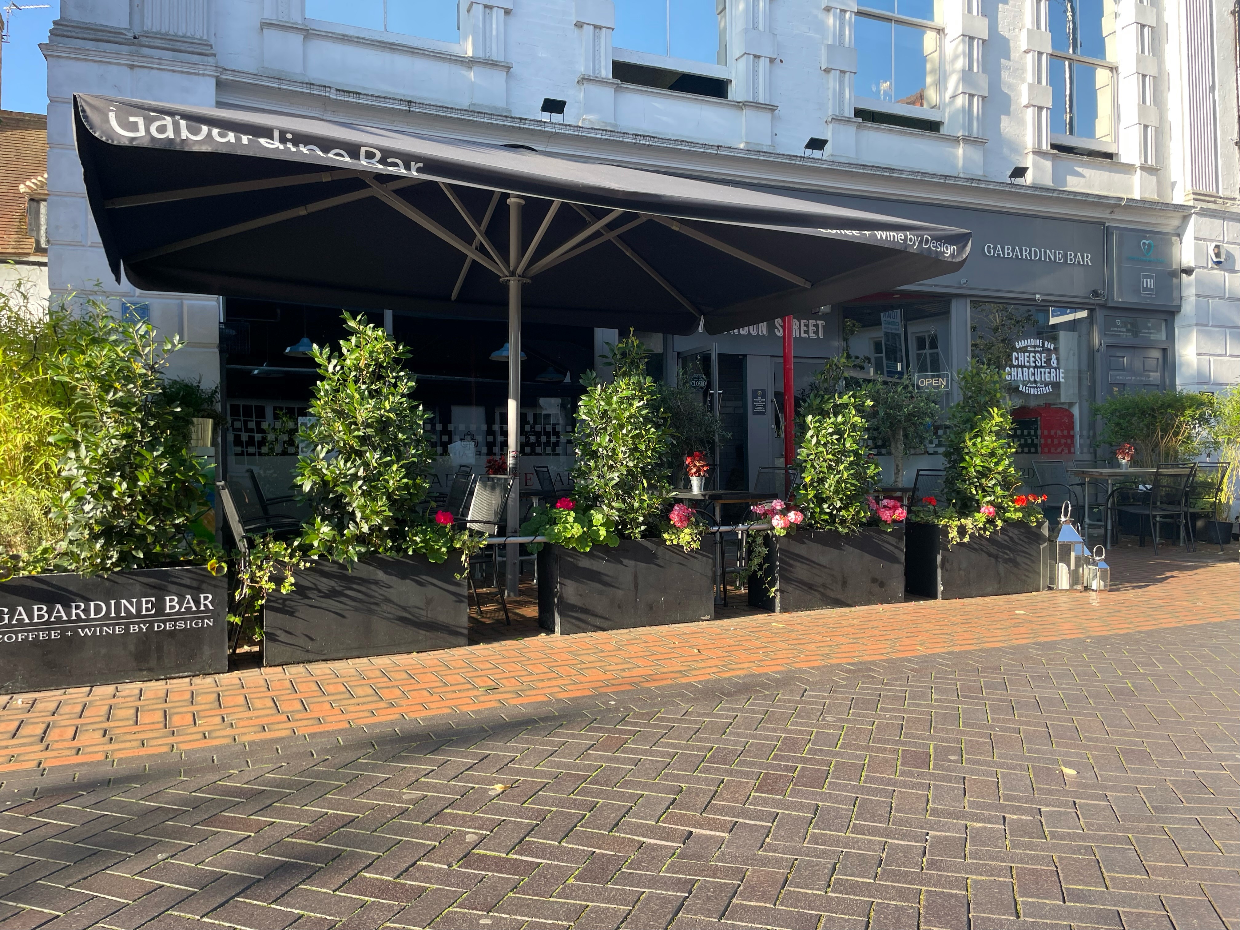 The outside of Gabardine, you can see planters with flowers in, a table with an umbrella up.