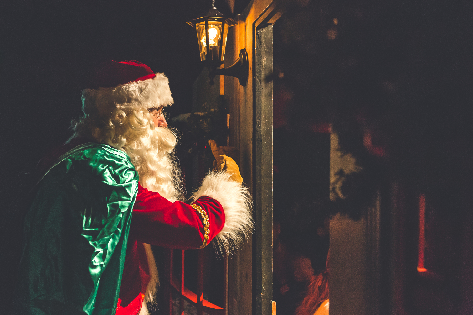 A nostalgic image shows Father Christmas behind a set at a Christmas show. The image is dark with dim atmospheric lighting. Santa holds a green sack over their shoulder.